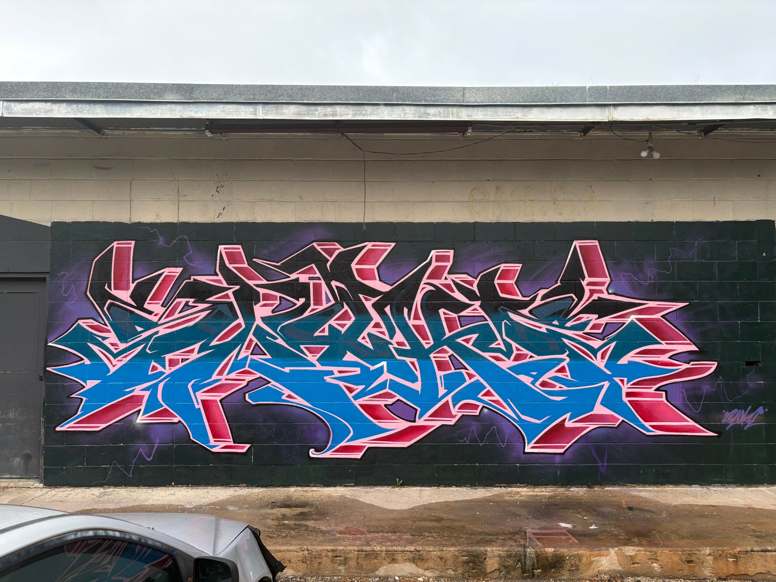 Colorful graffiti art with pink, purple, blue, and black elements on a wall, featuring abstract shapes and sharp lines.