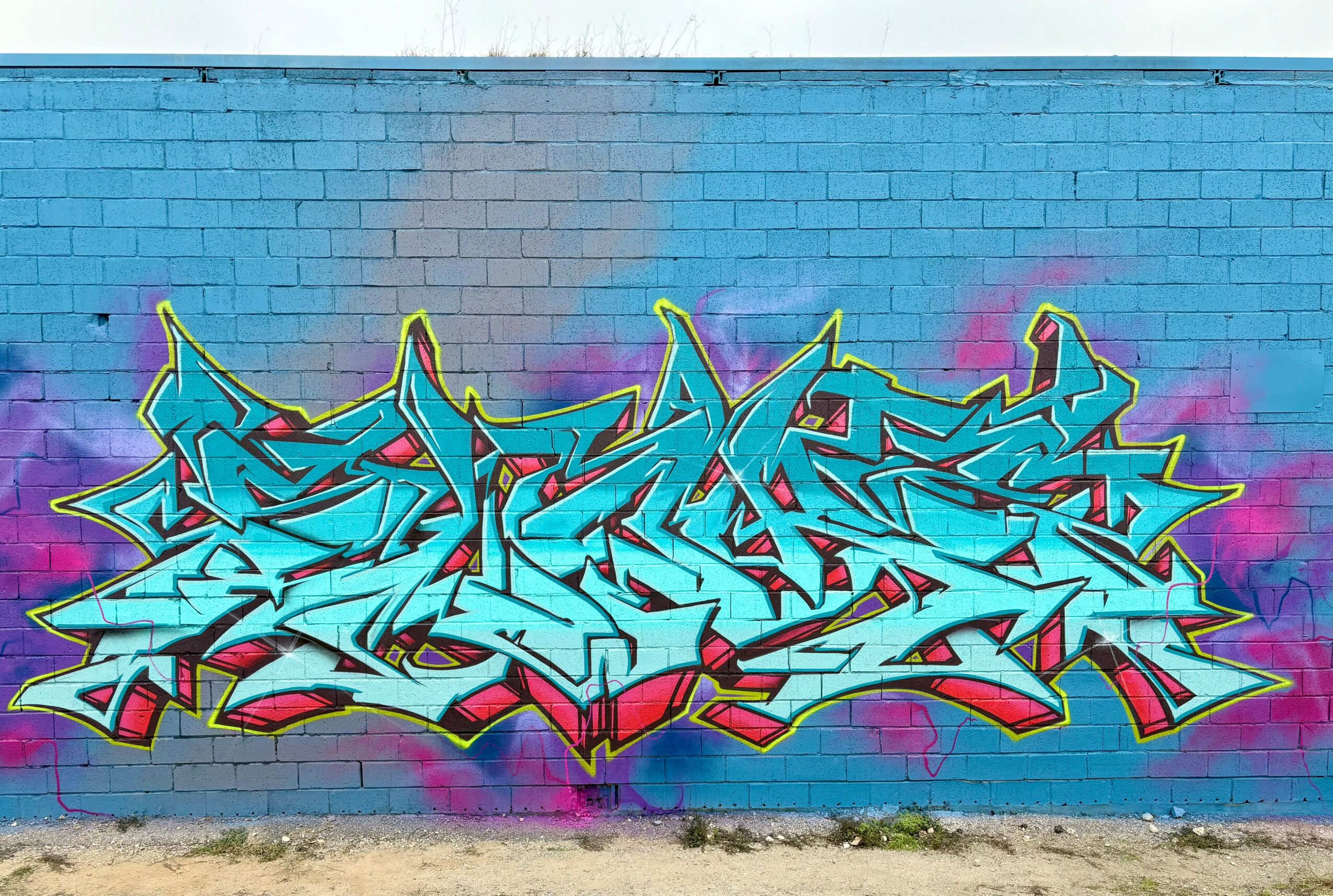 Bright blue graffiti with yellow outlines on a blue brick wall, featuring stylized, angular lettering with pink accents.