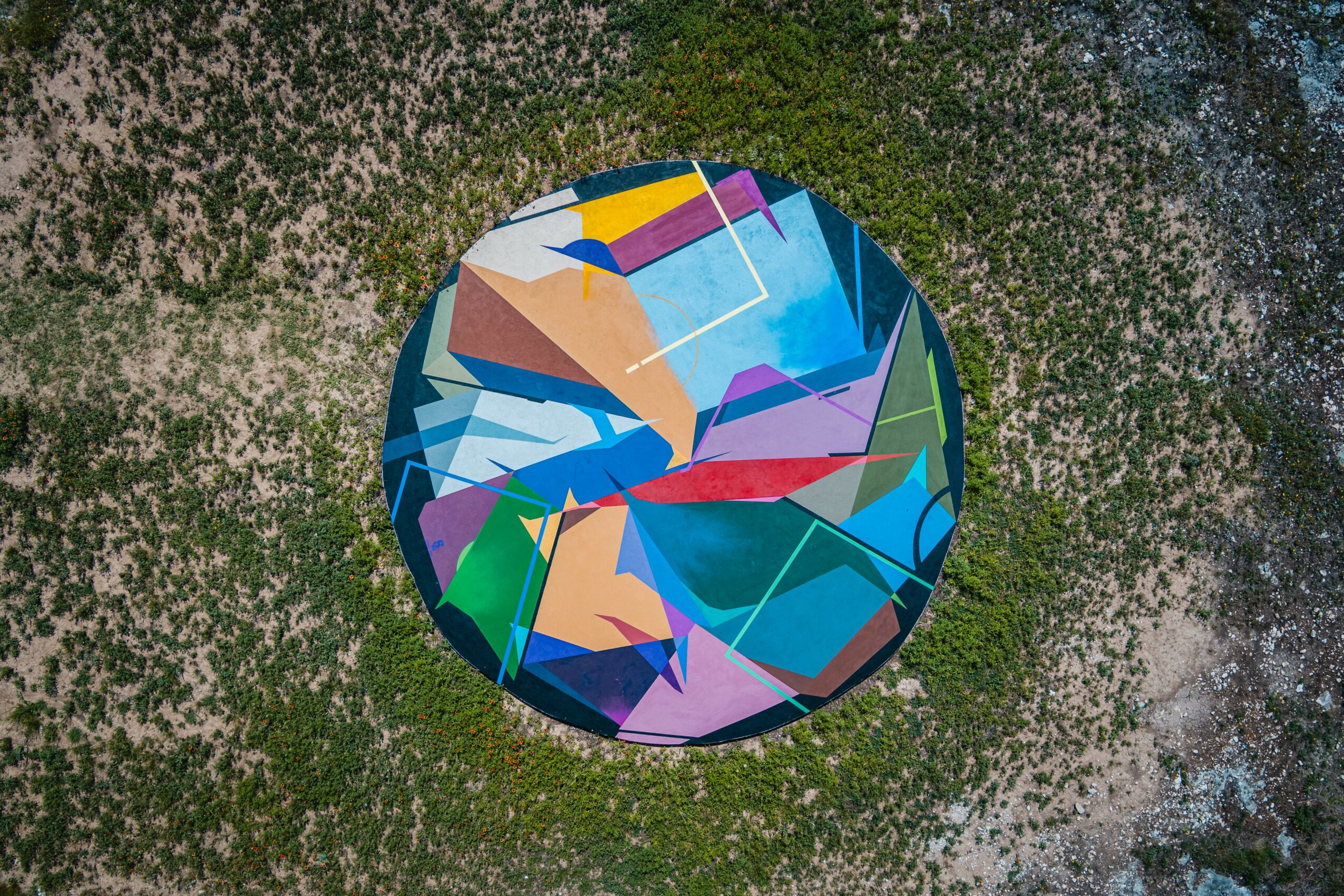 Colorful circular mural with abstract shapes on a wall, surrounded by moss and dirt ground