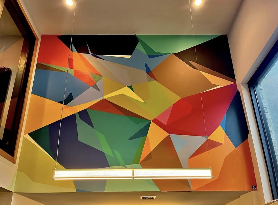 Colorful abstract geometric mural on wall with ceiling lights and window, vibrant shapes and colors.