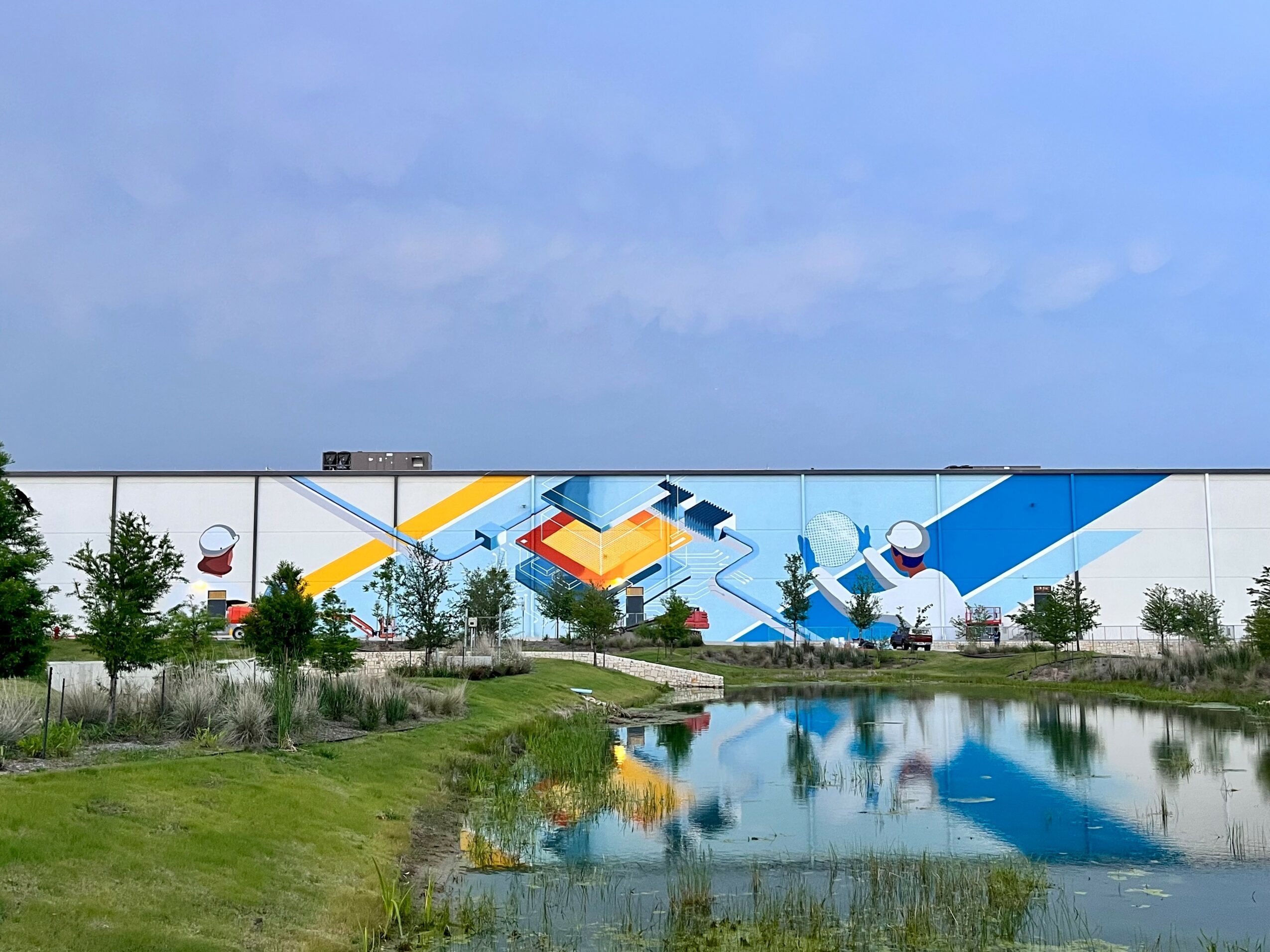 Mural on building wall depicts abstract geometric shapes and a person in a white helmet, with a pond and trees in foreground.
