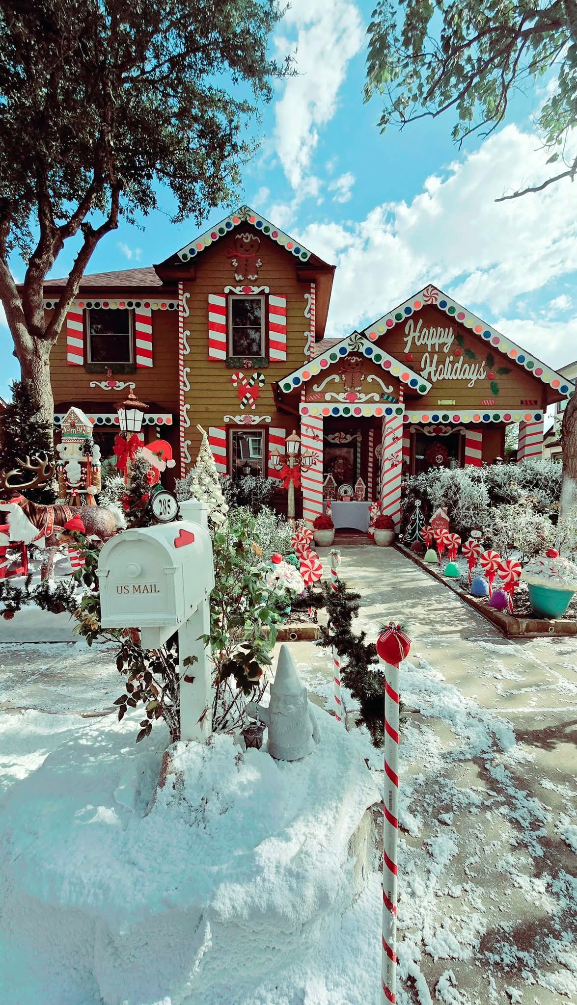 Two decorated houses with Christmas decorations, snow-covered yard, mailbox, and a pathway, under a blue sky with clouds.