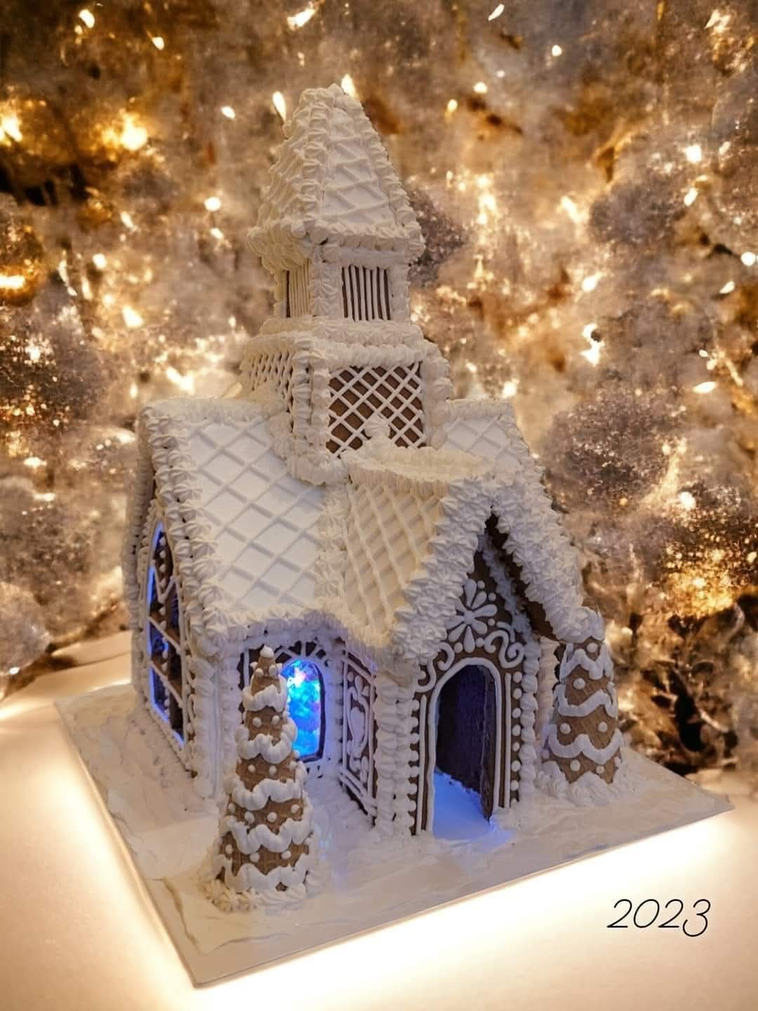 White gingerbread house with intricate details, illuminated with blue light, set against a sparkling background, with the year 2023 in the corner.