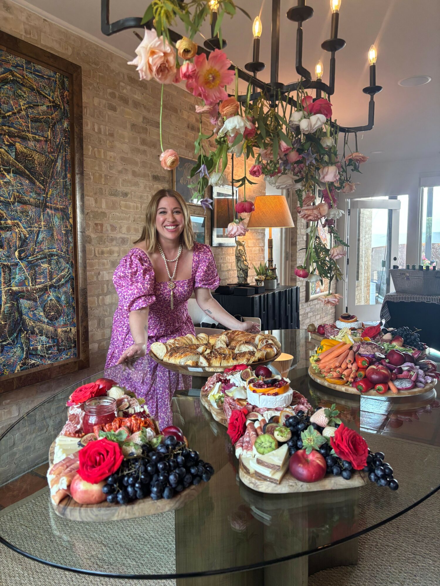 Woman in pink dress sitting at table with fruit and desserts, flowers hanging above, in a room with brick wall and window.