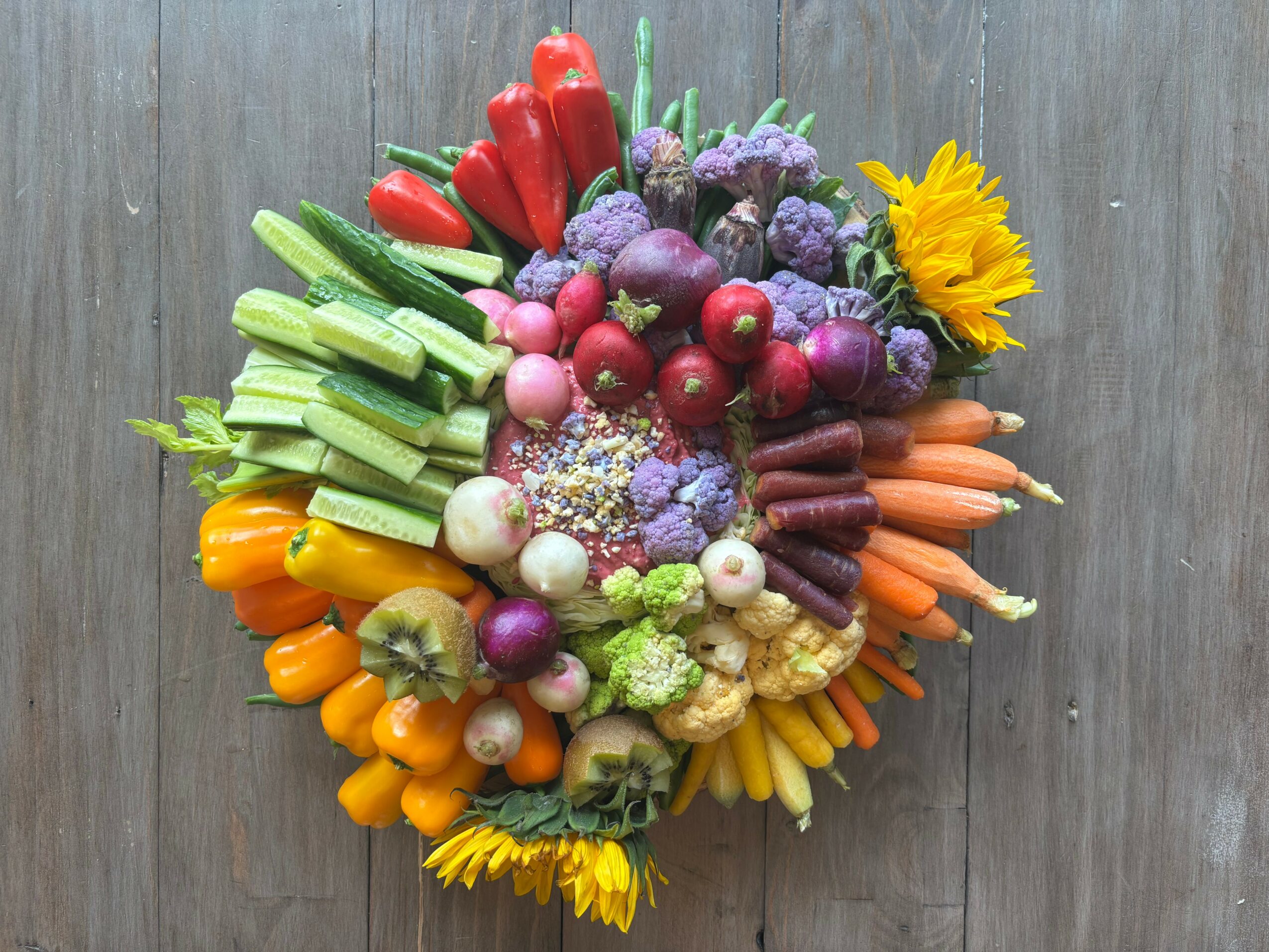 Colorful vegetable arrangement with red, green, orange, purple, and yellow vegetables and a sunflower, on a wooden surface.