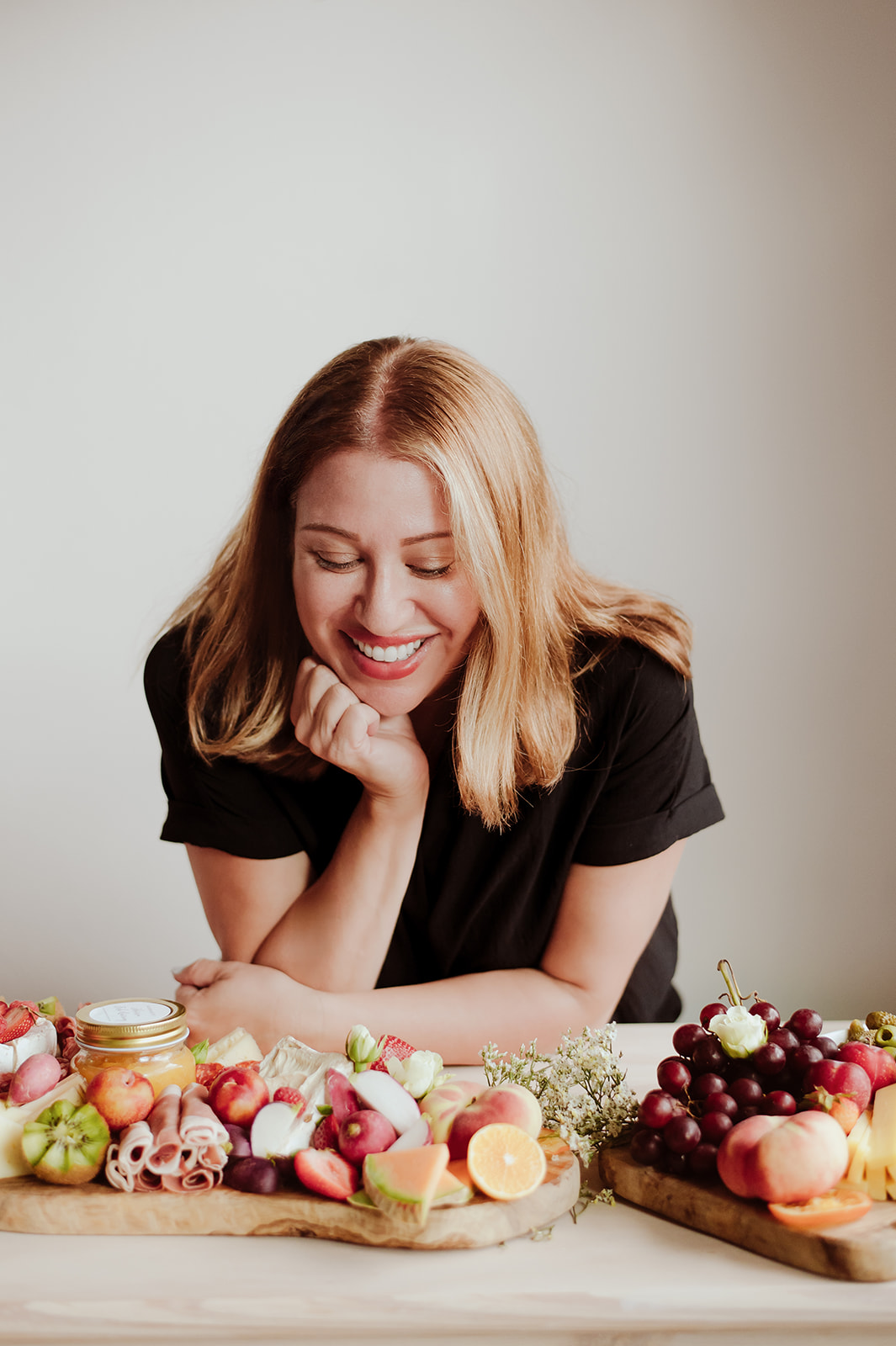 Smiling woman with blonde hair resting her chin on her hand, surrounded by colorful desserts on a table.