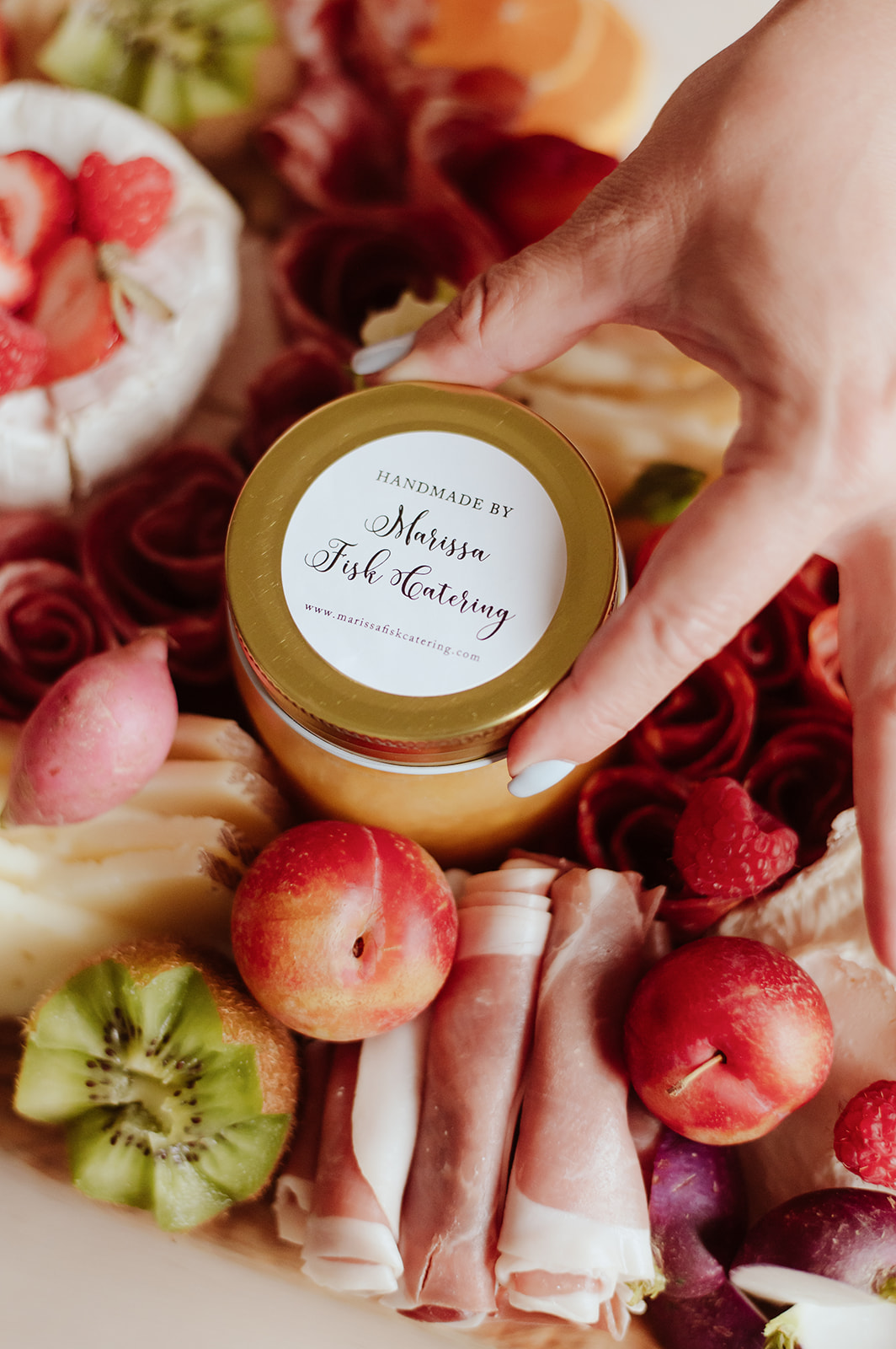 Hand holding a jar labeled 'Handmade by Mom's 3rd Morning' surrounded by strawberries, kiwi, grapes, and rolled slices of meat or cheese.
