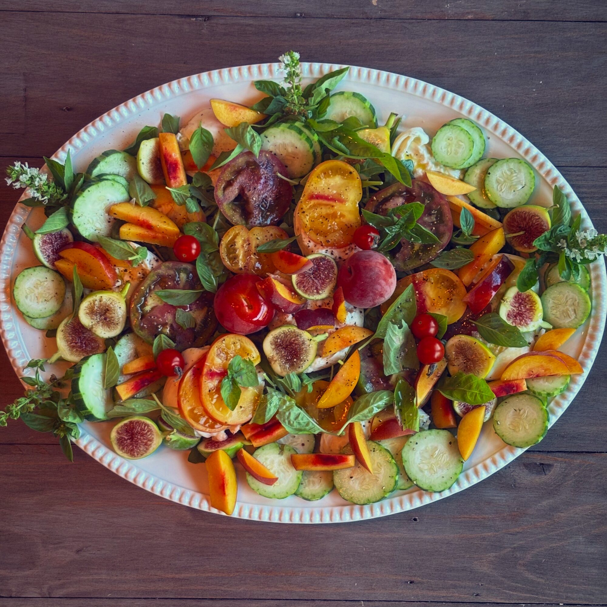 Colorful fruit and vegetable salad on a white oval platter, including grapes, cherry tomatoes, cucumber, and sliced peaches, on a dark wooden surface.