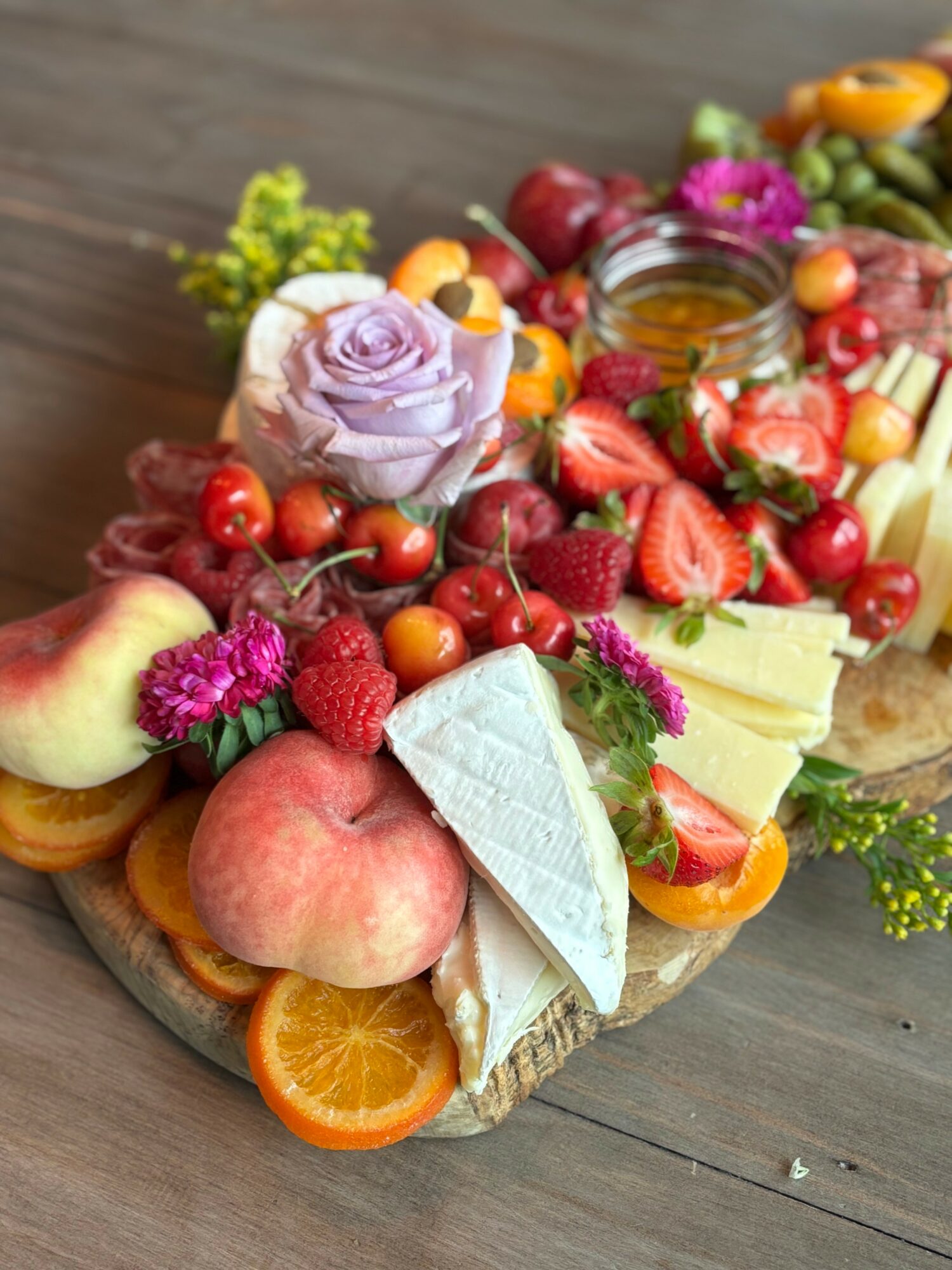 Cheese, strawberries, peaches, grapes, and flowers arranged on a wooden board with a jar of honey in the center.