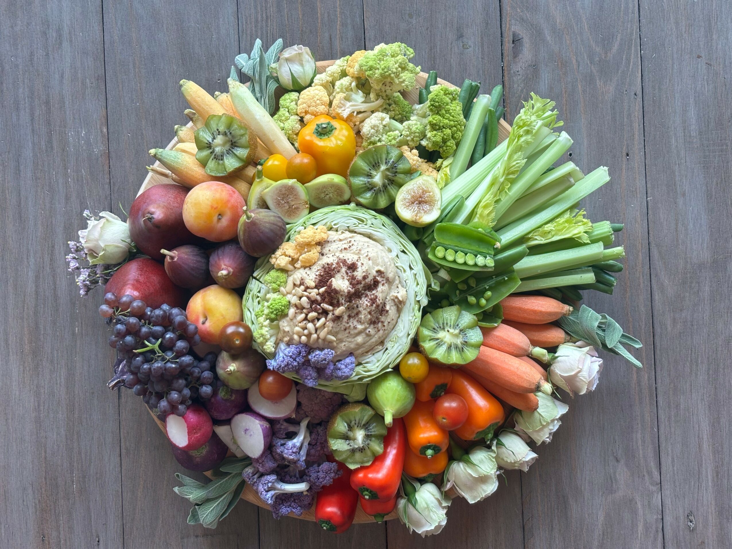 Assorted fresh vegetables and fruits arranged on a wooden surface, including tomatoes, carrots, celery, grapes, and apples.