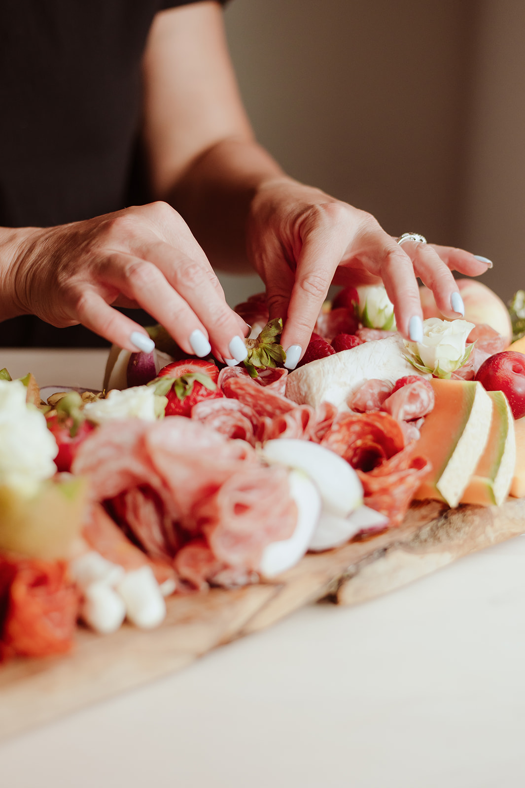 Person's hands arranging food on a wooden board with meats, cheeses, and vegetables, in a close-up view.