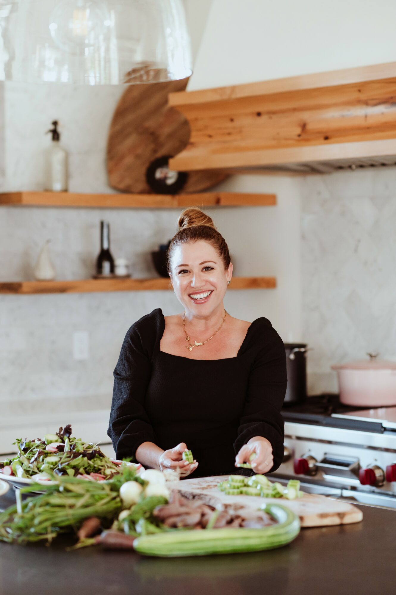 Woman with red hair in a bun smiling while preparing food in a kitchen with wooden shelves and a stove
