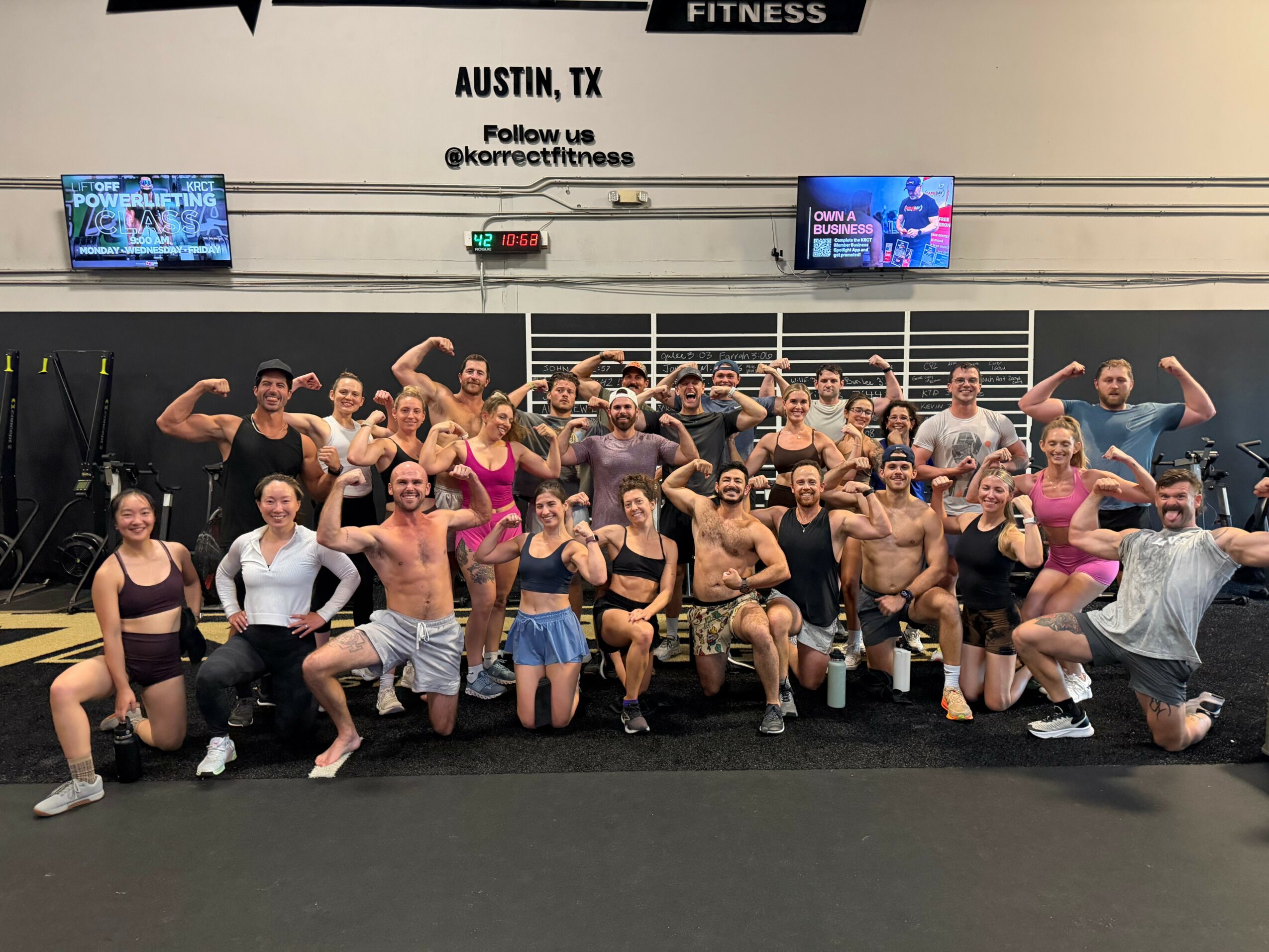 Group of people in workout attire posing in a gym, some flexing muscles, with two screens and a sign reading Austin, TX.