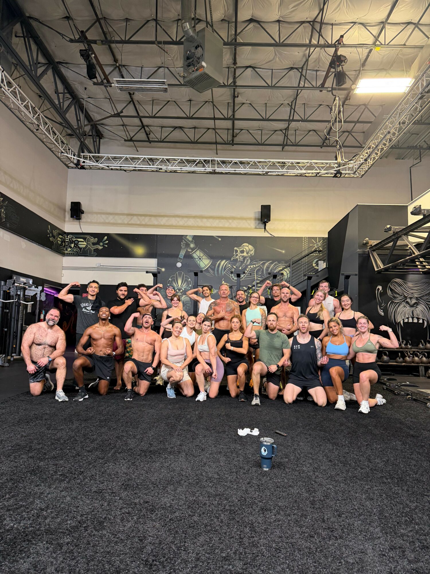 Group of people posing on stage with a black backdrop, some holding drinks, in an indoor event space.