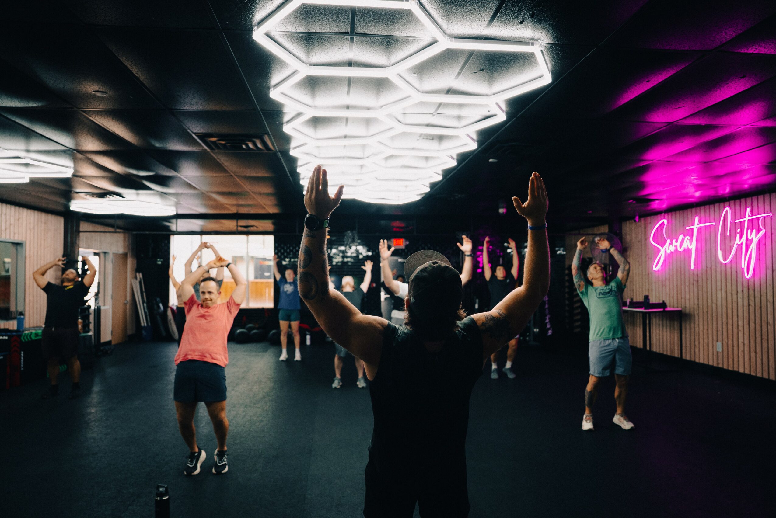 People participating in a dance class with a person leading, illuminated ceiling lights, and a neon sign reading 'Sweet City'.