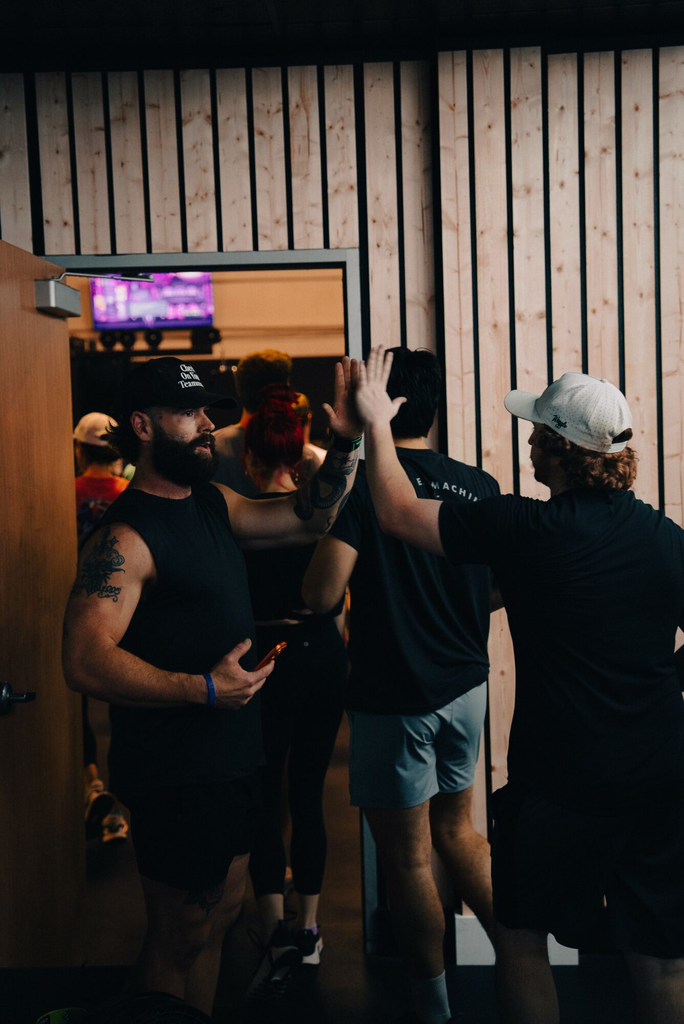 Three people high-fiving in a room with wooden wall panels and a mirror, one holding a phone, casual attire.
