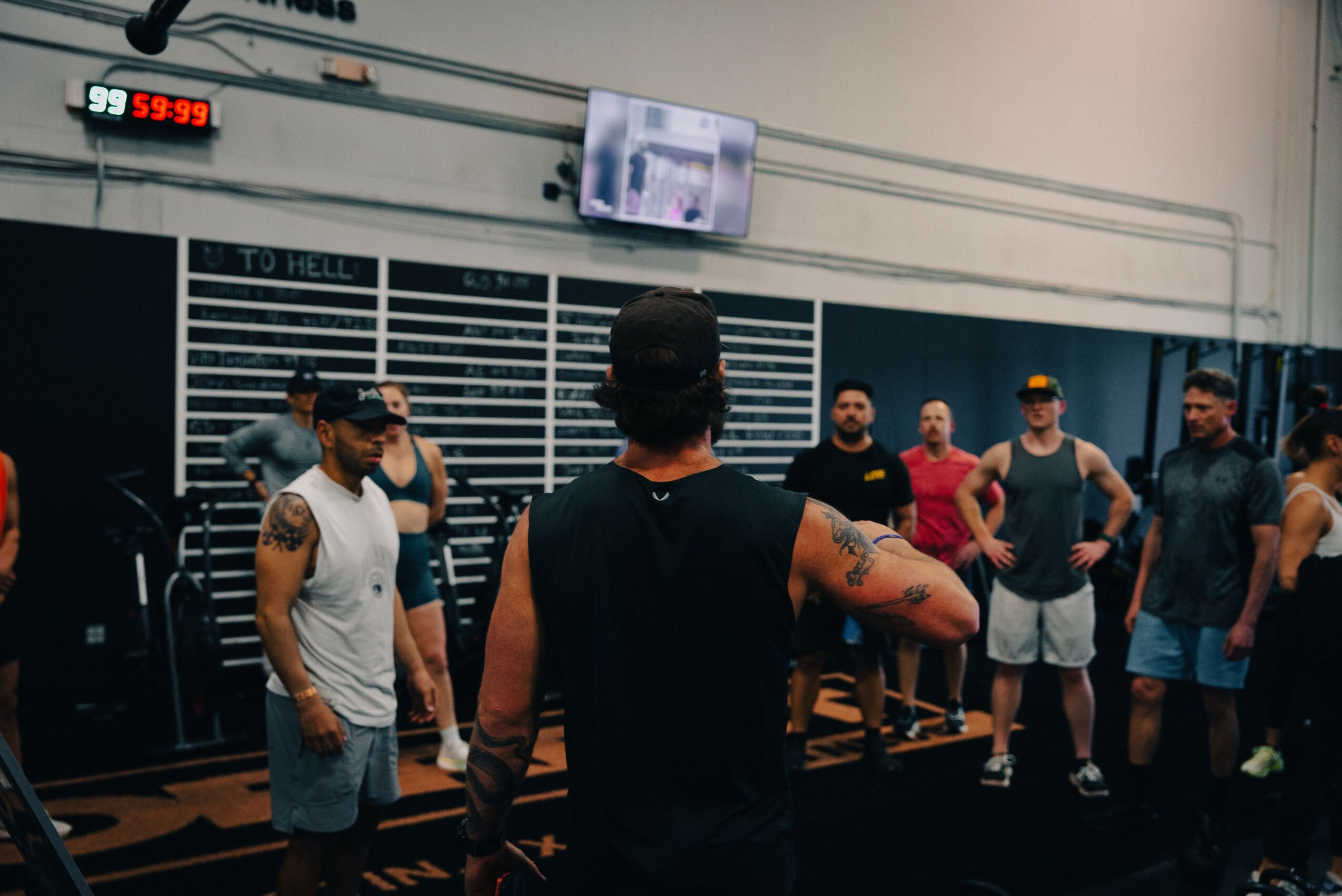 Group of people standing in a gym, some with tattoos, facing a person in the center, with a digital clock and TV screen above.