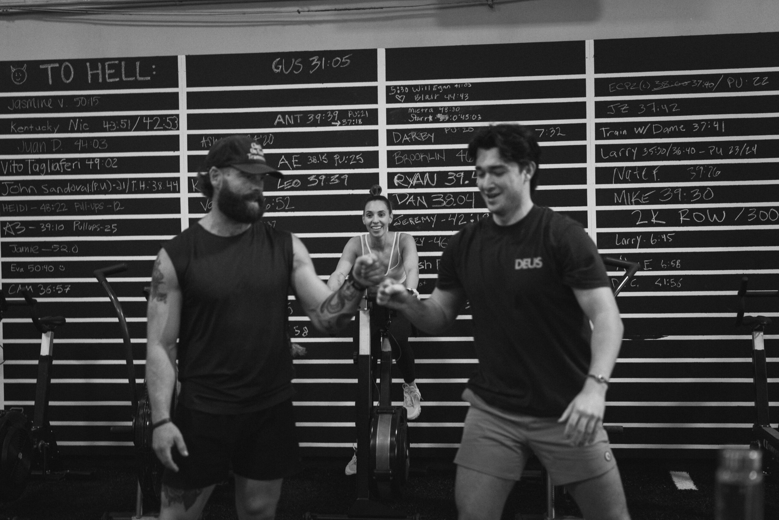 Two men shake hands in front of a blackboard with writing, a woman stands behind them, smiling, in a gym or fitness setting.