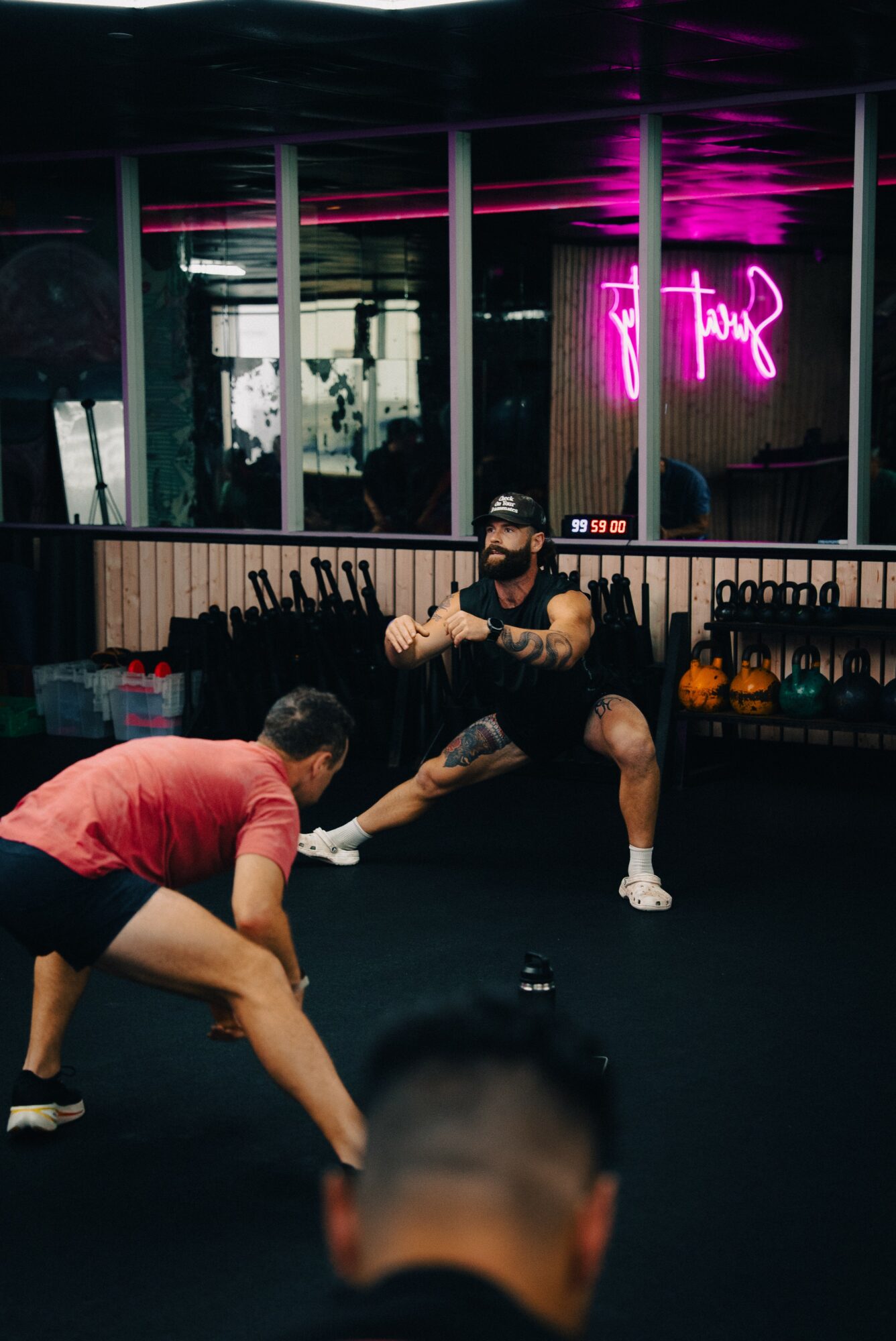 Person in black workout clothes and cap demonstrating a squat in a gym with kettlebells and neon pink sign, others watching.
