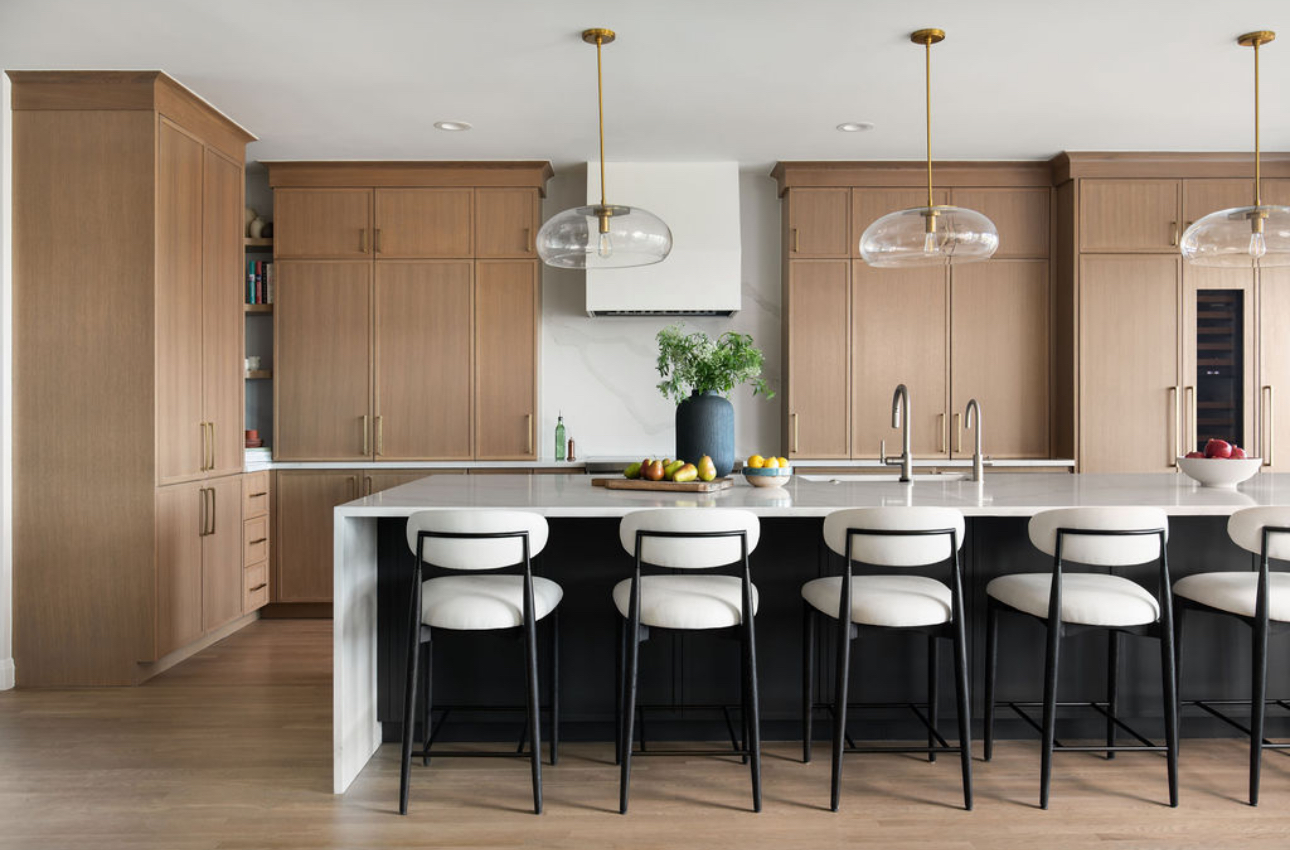 Modern kitchen with wooden cabinets, white island, six white and black bar stools, pendant lights, and a potted plant.