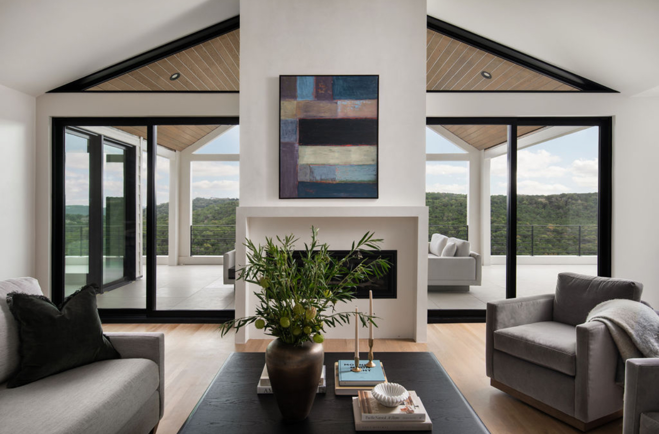 Living room with large windows, fireplace, and modern furniture, overlooking a green landscape outside.