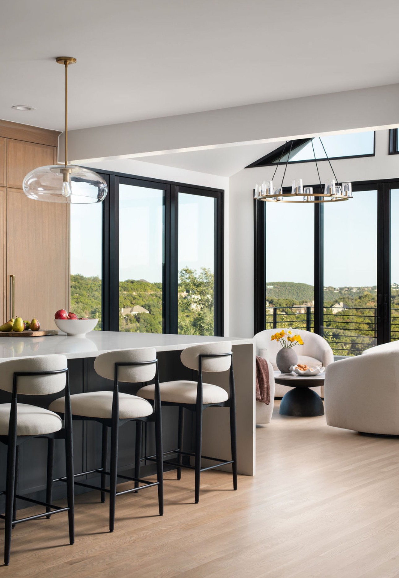 Modern kitchen and living area with large windows, bar stools, and contemporary lighting fixtures, bright and airy space.