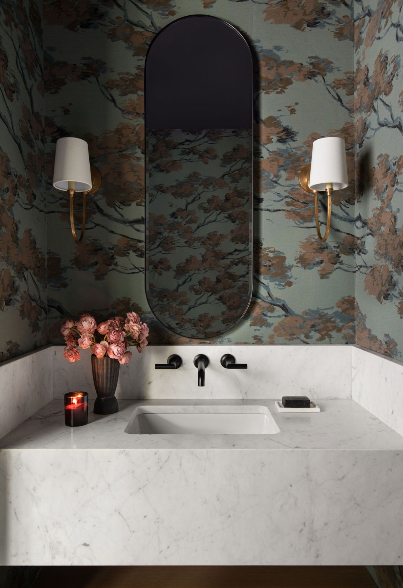 Bathroom sink with mirror, floral wallpaper, two wall-mounted lights, pink flowers, candle, and soap dispenser.