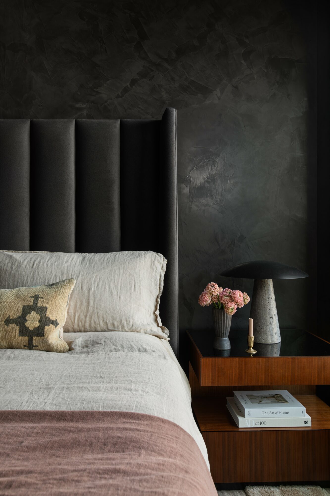 Part of a bedroom with a bed, headboard, nightstand, and decorative items against a dark wall.