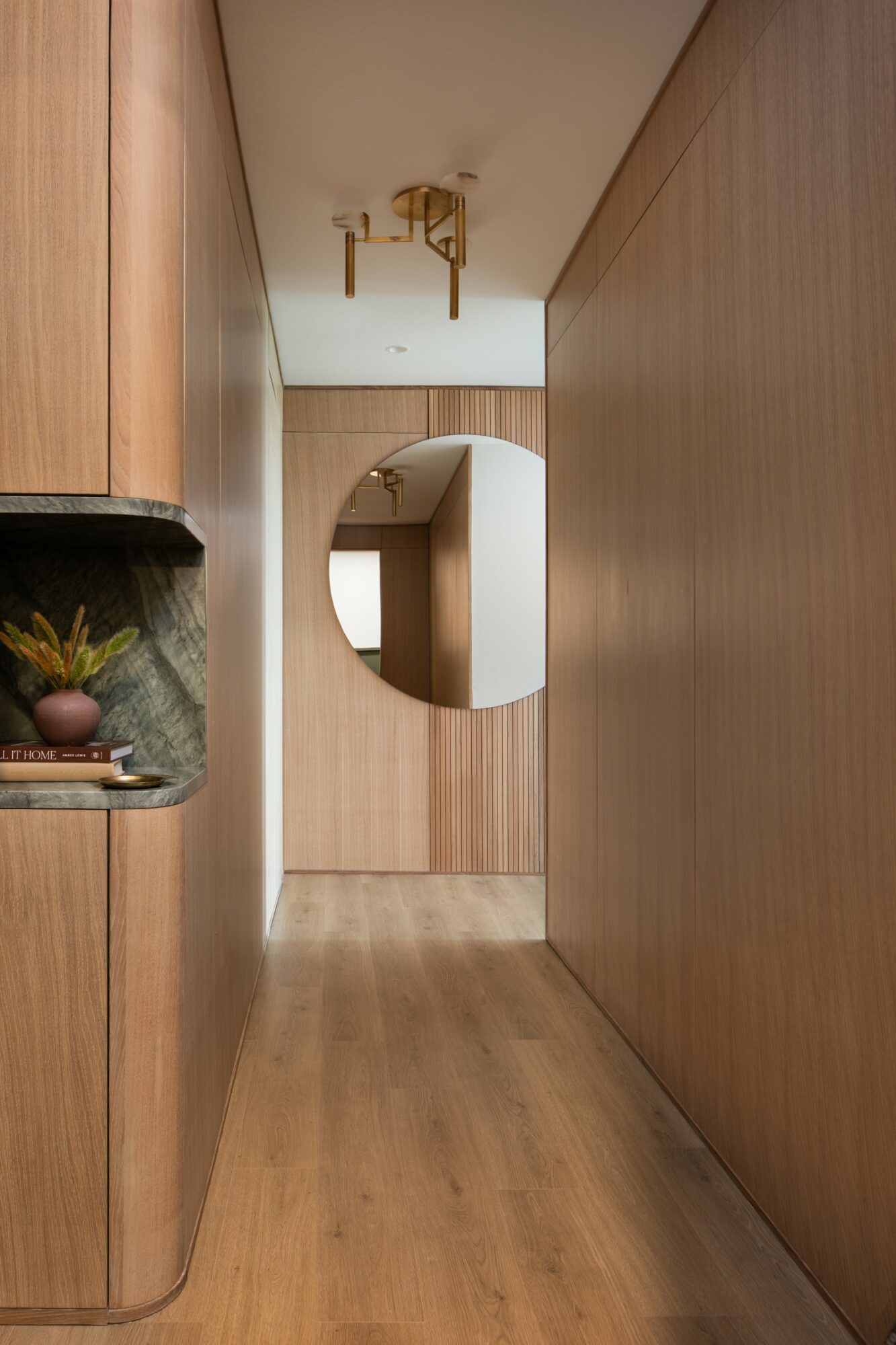 Narrow hallway with wooden walls and floor, mirror on the back wall, ceiling light fixture, and a small shelf with plants on the left.