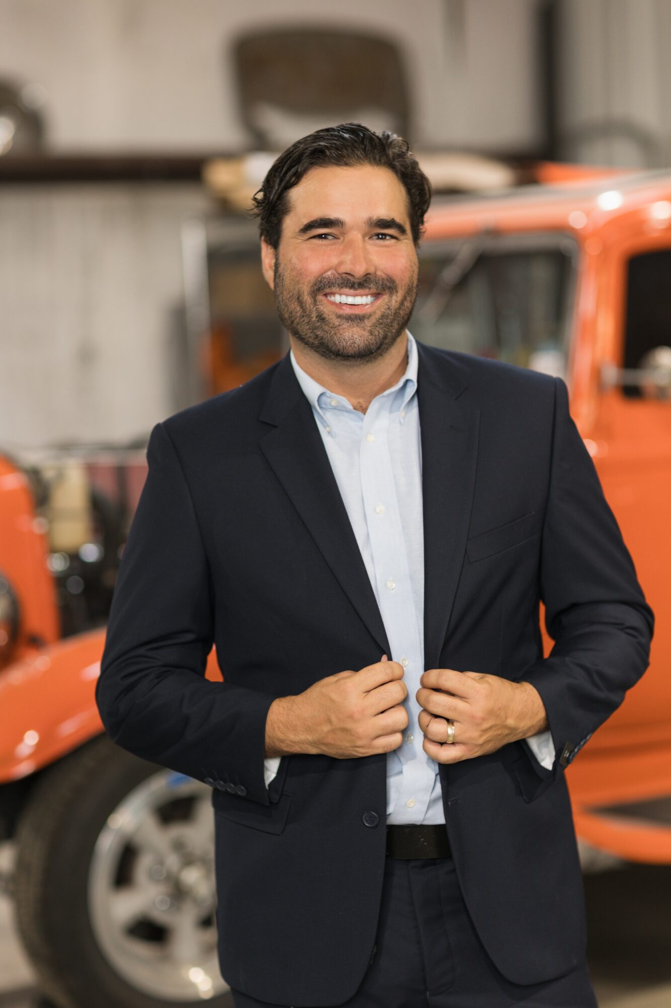 Man in a suit smiling, standing outdoors with an orange vehicle in the background.