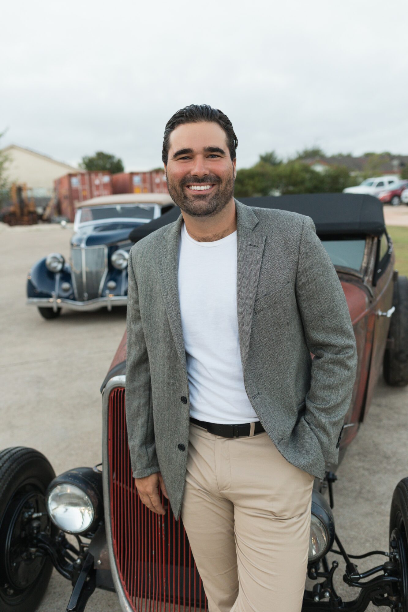 Man with dark hair and beard smiling, wearing a gray blazer and white shirt, standing outdoors near vintage cars.