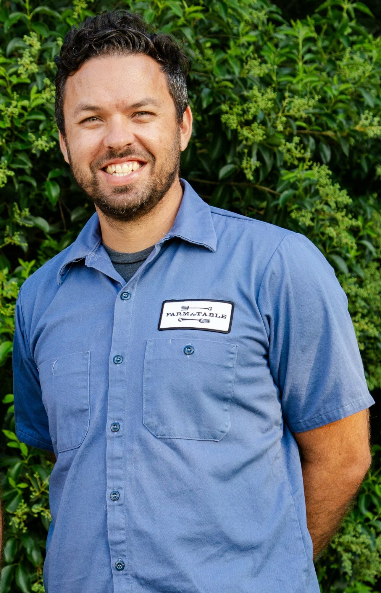 Man with dark hair and beard smiling outdoors, wearing a blue work shirt with a name tag, green foliage background.