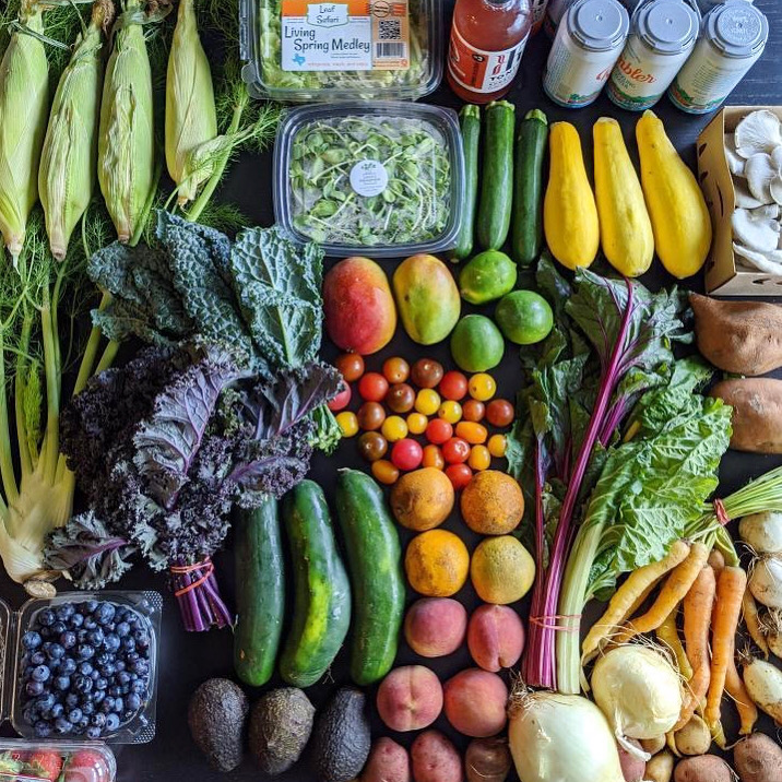 Assorted fresh vegetables and fruits including corn, lettuce, cucumbers, tomatoes, apples, oranges, carrots, and blueberries.
