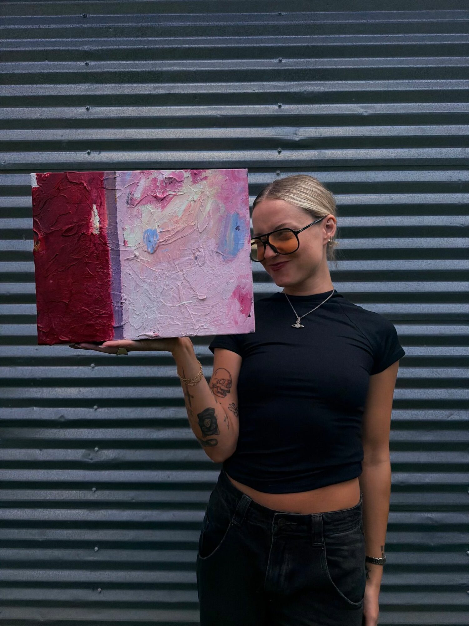 Person with blonde hair, glasses, black shirt, and tattoos holding a pink and red painting against a striped background.