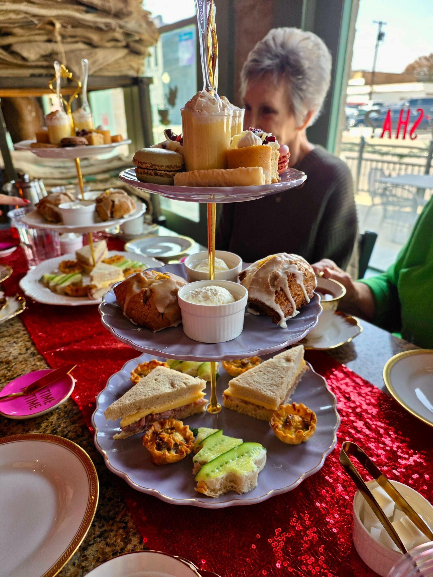 A woman with short gray hair sits at a table with a three-tiered dessert stand and various pastries, in a bright setting.