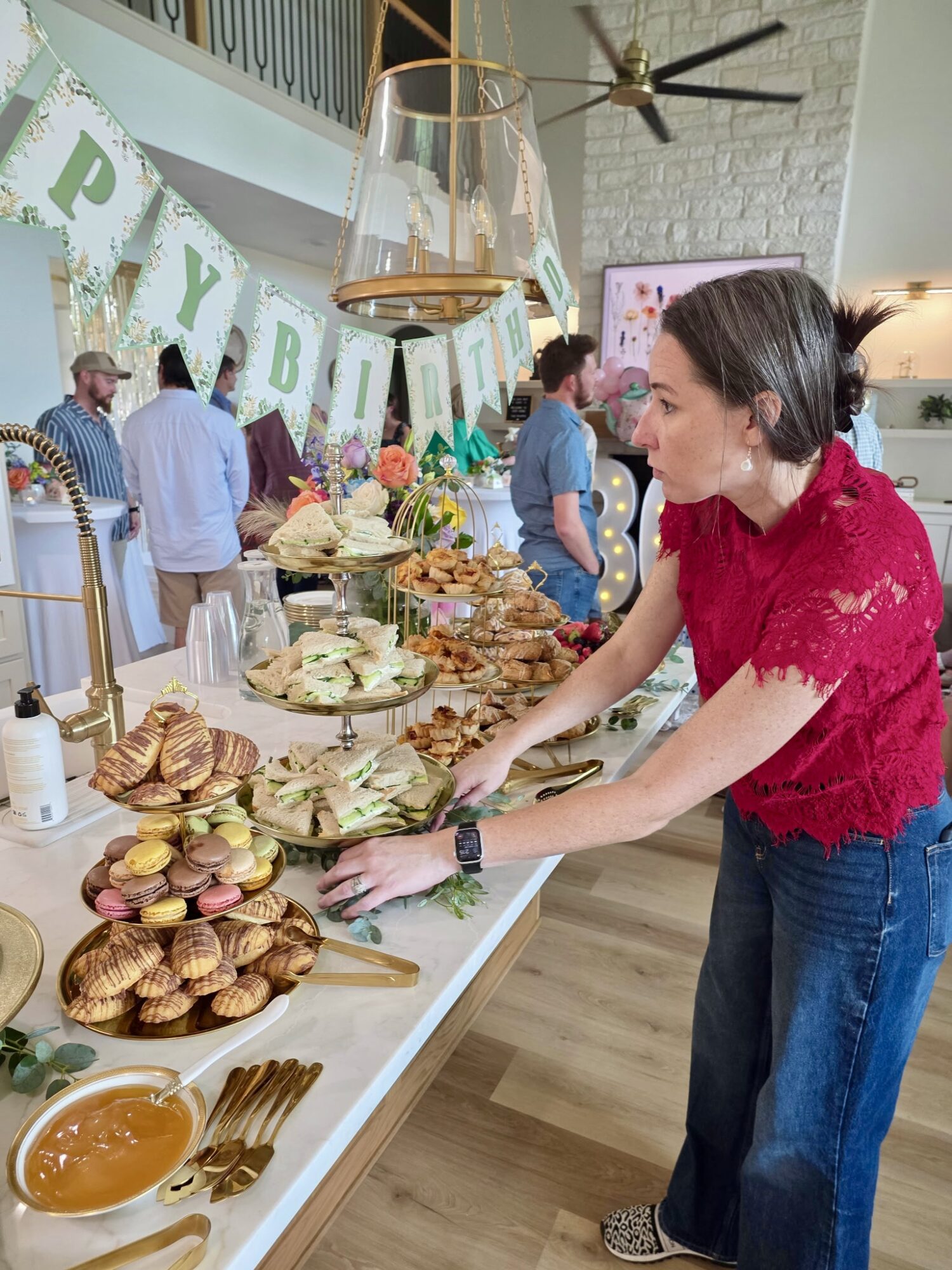 Woman in red top and jeans reaching for food at a decorated party table with desserts and decorations.
