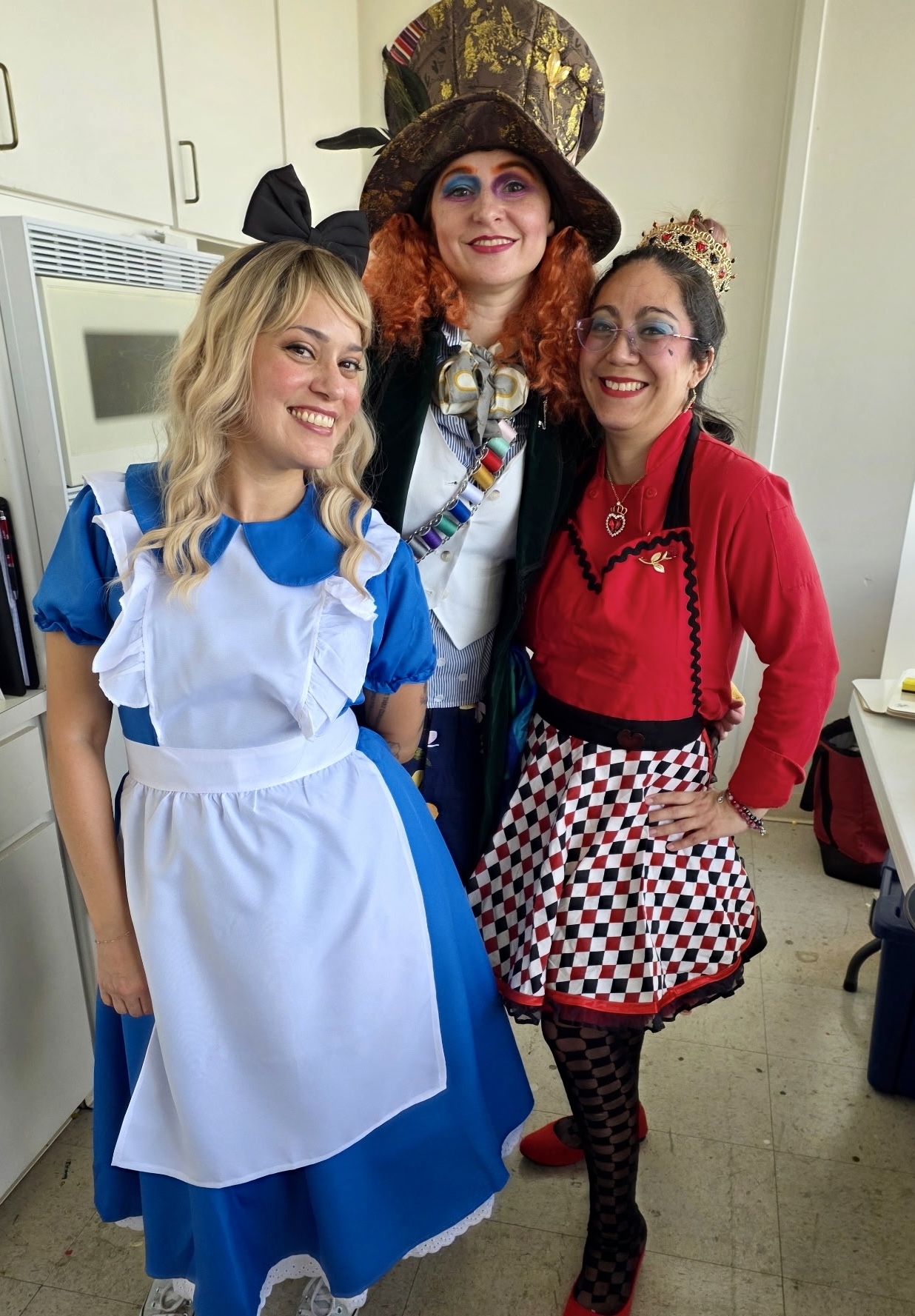 Three women in costumes smiling indoors, one dressed as Alice in Wonderland, another as a queen, and the third in a red outfit with a checkered apron.