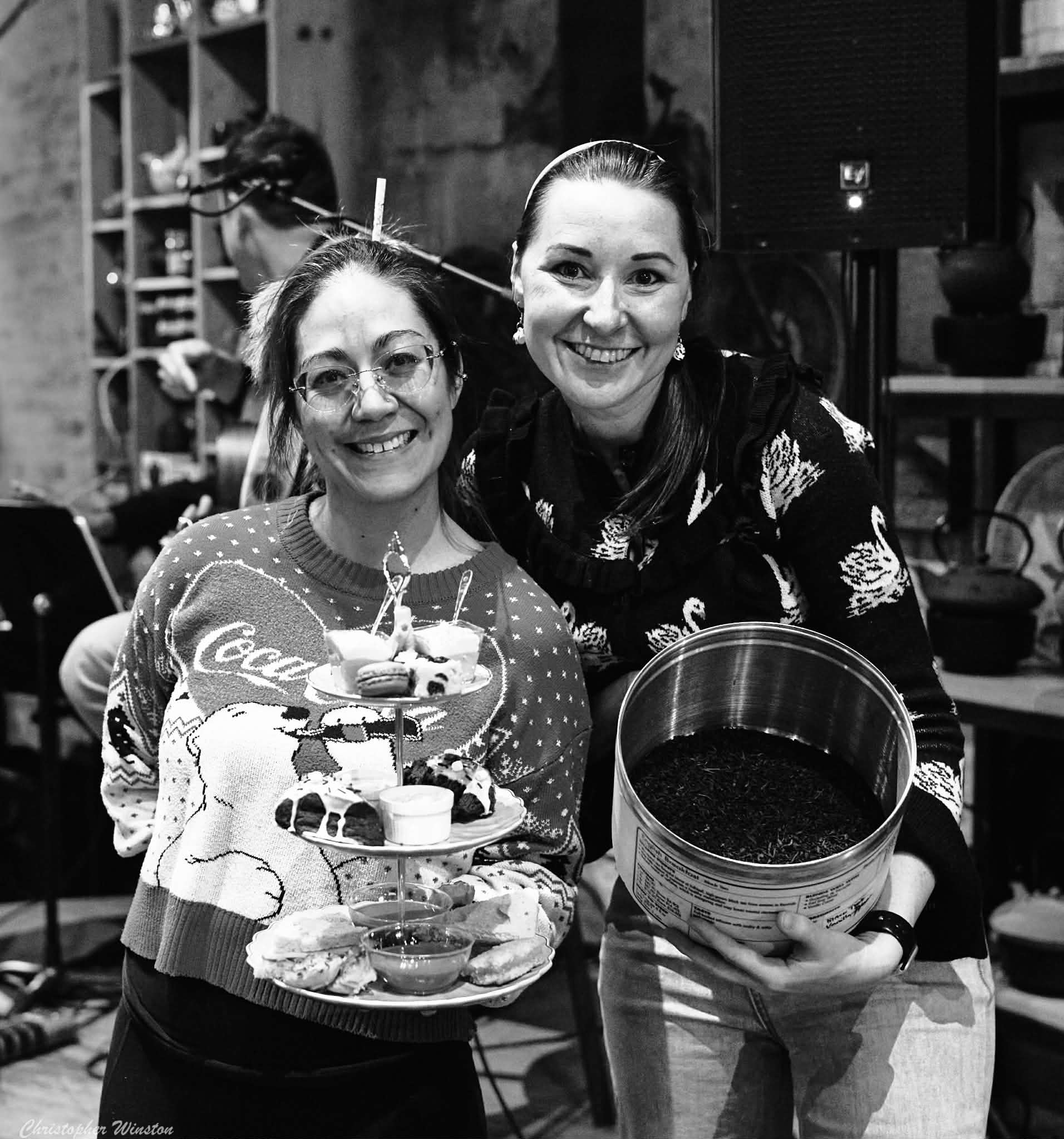 Two women smiling, one holding a multi-tiered tray of desserts, in a cozy indoor setting.