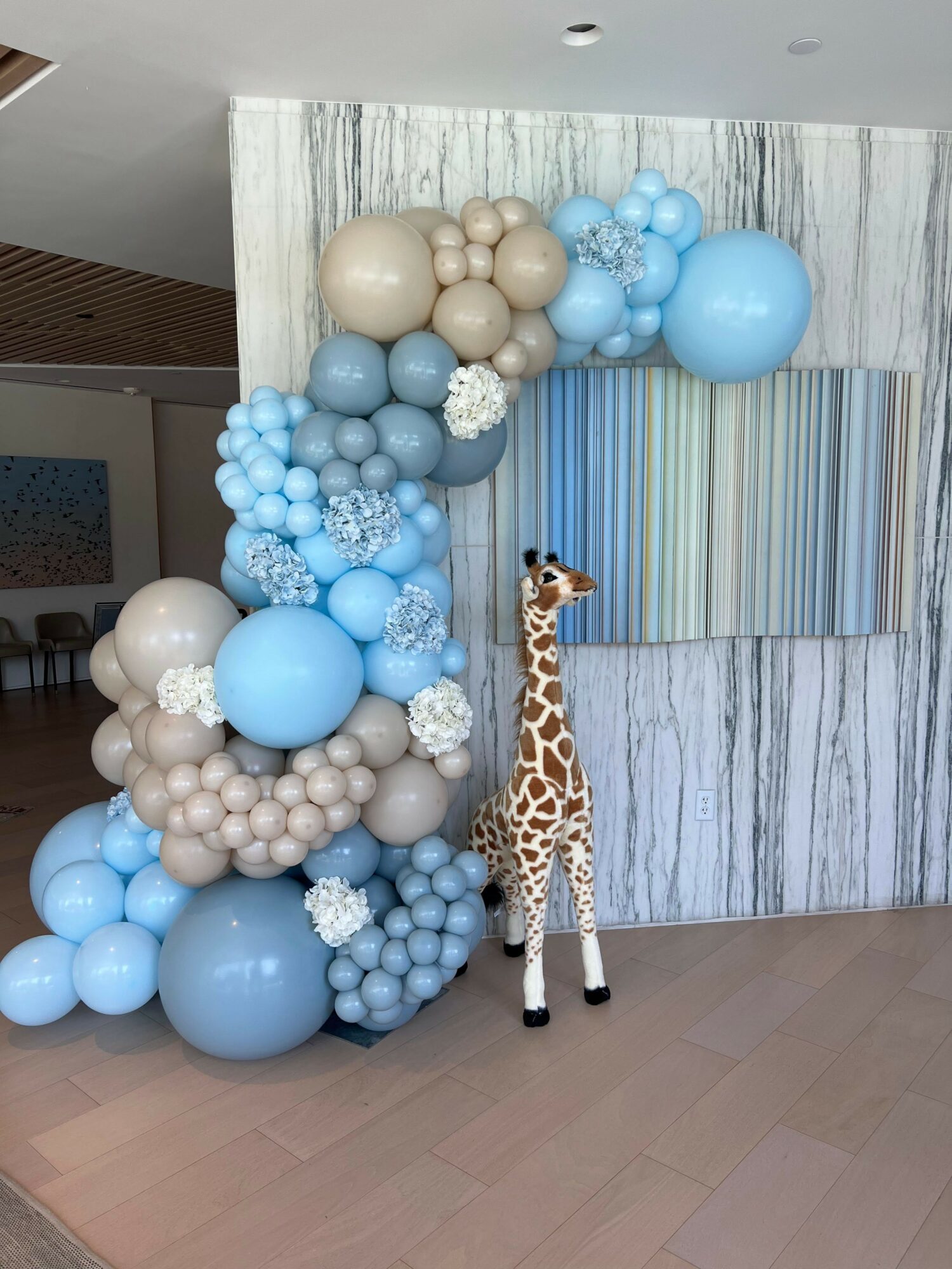 Giraffe figurine surrounded by blue, beige, and white balloons on a wooden floor and marble wall background.