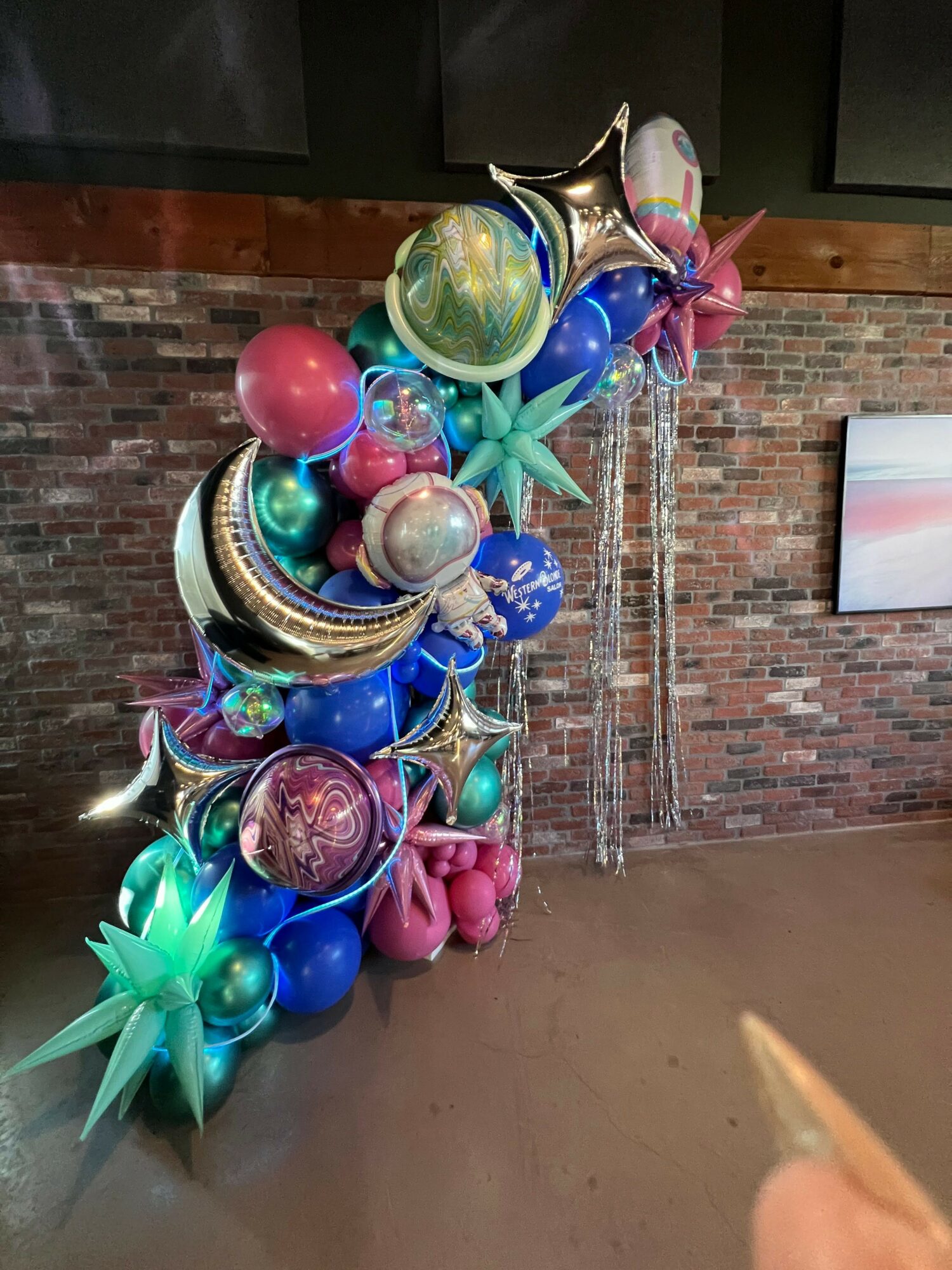 Balloon arrangement with stars, planets, and a rainbow, set against a brick wall background.
