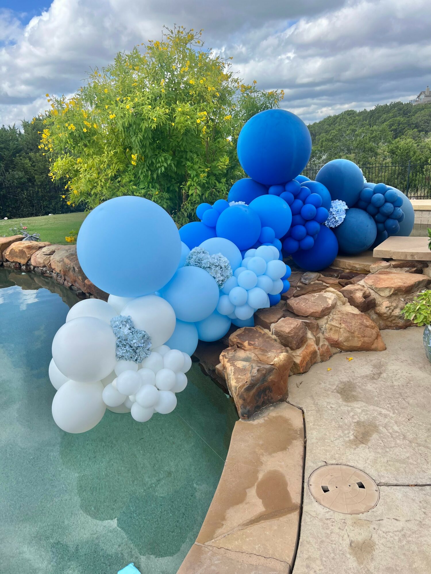 Colorful balloon arrangement with blue and white balloons on rocks near water, outdoors with trees and cloudy sky.