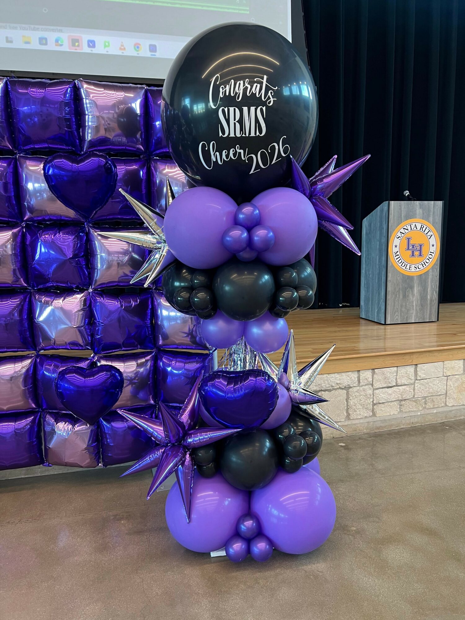 Balloon arrangement with purple, black, and silver balloons, including a large black balloon with white text, on a beige floor.