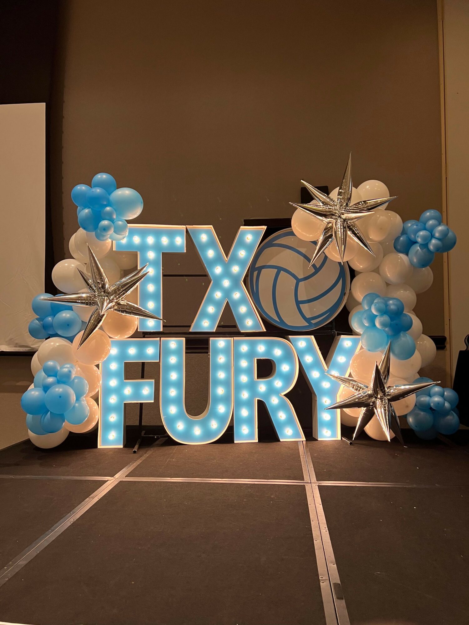 Decorative sign with balloons and pinwheels, spelling 'EX' and 'US' in illuminated letters, on a tiled floor.