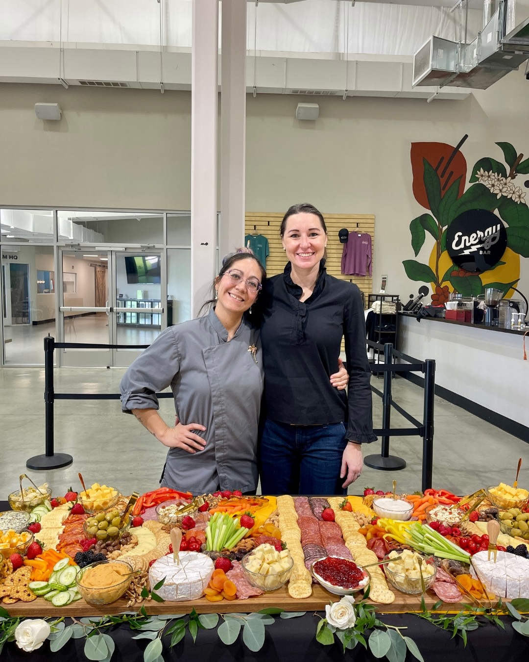 Two women stand behind a table with various food items, smiling indoors with a colorful mural in the background.