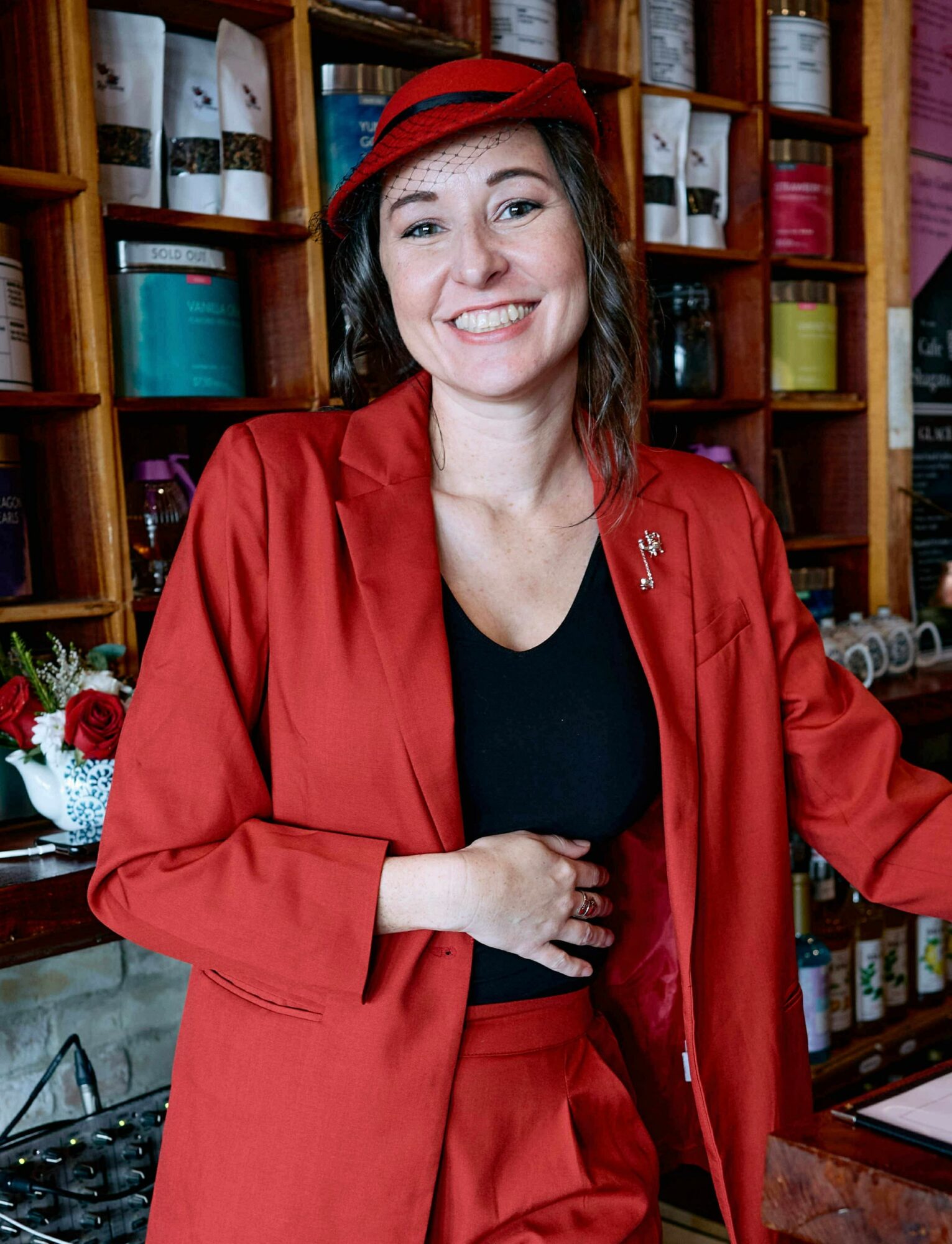 Woman smiling in red blazer and hat, standing in front of colorful shelves, holding her hand near her waist.
