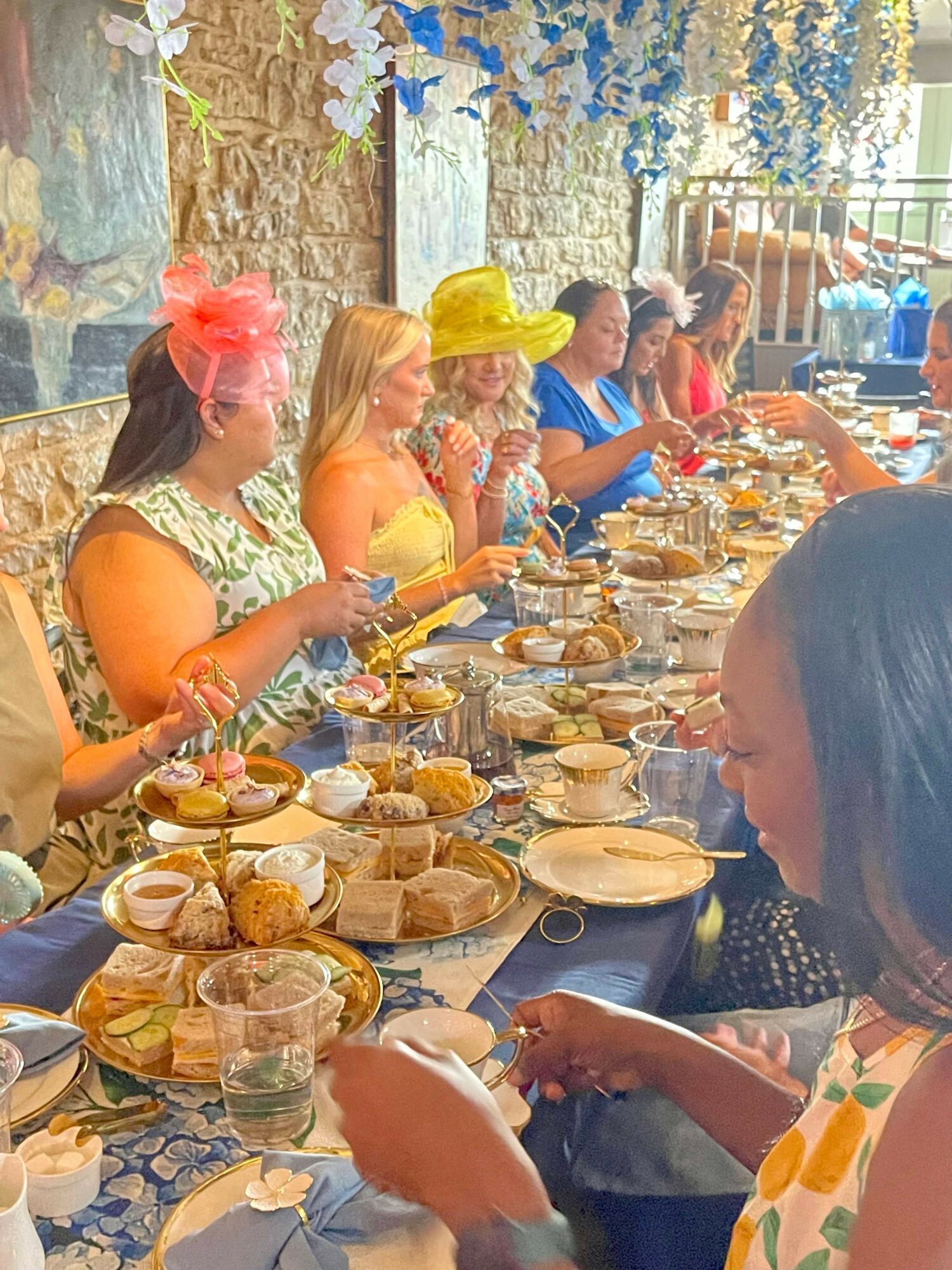 People sitting at a long table with food, drinks, and decorations, enjoying a gathering outdoors under a canopy with flowers.