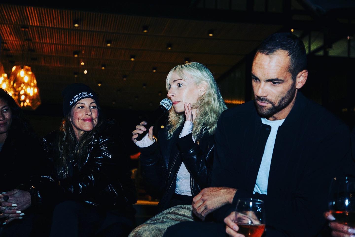 Group of people at an indoor event, one woman speaking into a microphone, others listening, dim lighting, warm ambiance.