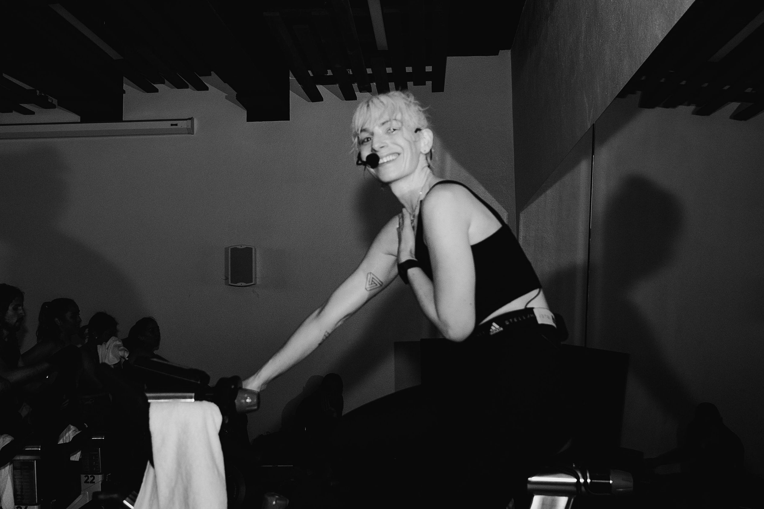 Person with short, curly hair smiling and sitting on a chair, holding a microphone, in a dimly lit room with an audience.