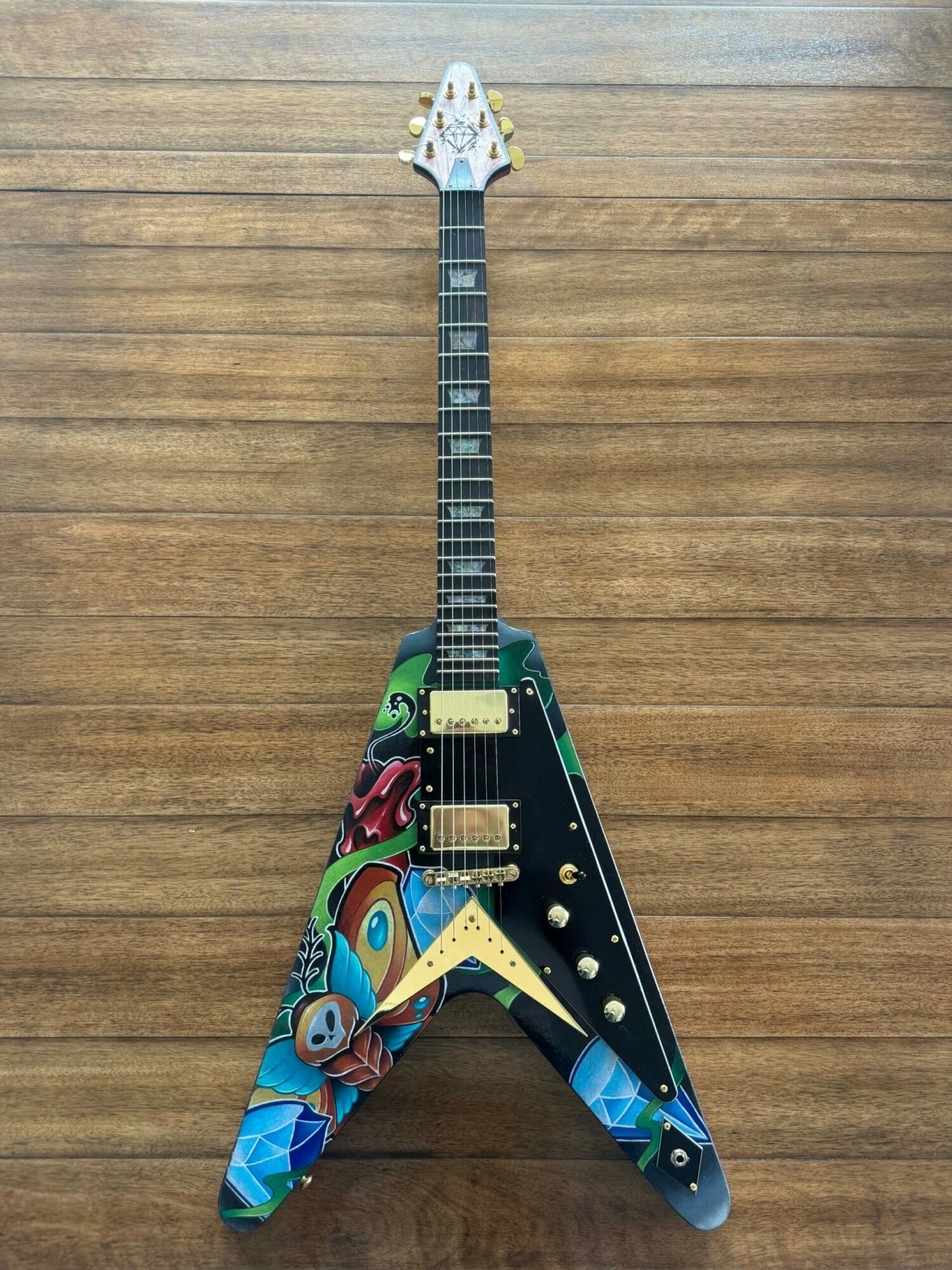 Electric guitar with a colorful, abstract painted design, lying on a wooden floor, with a V-shaped body and a long neck.