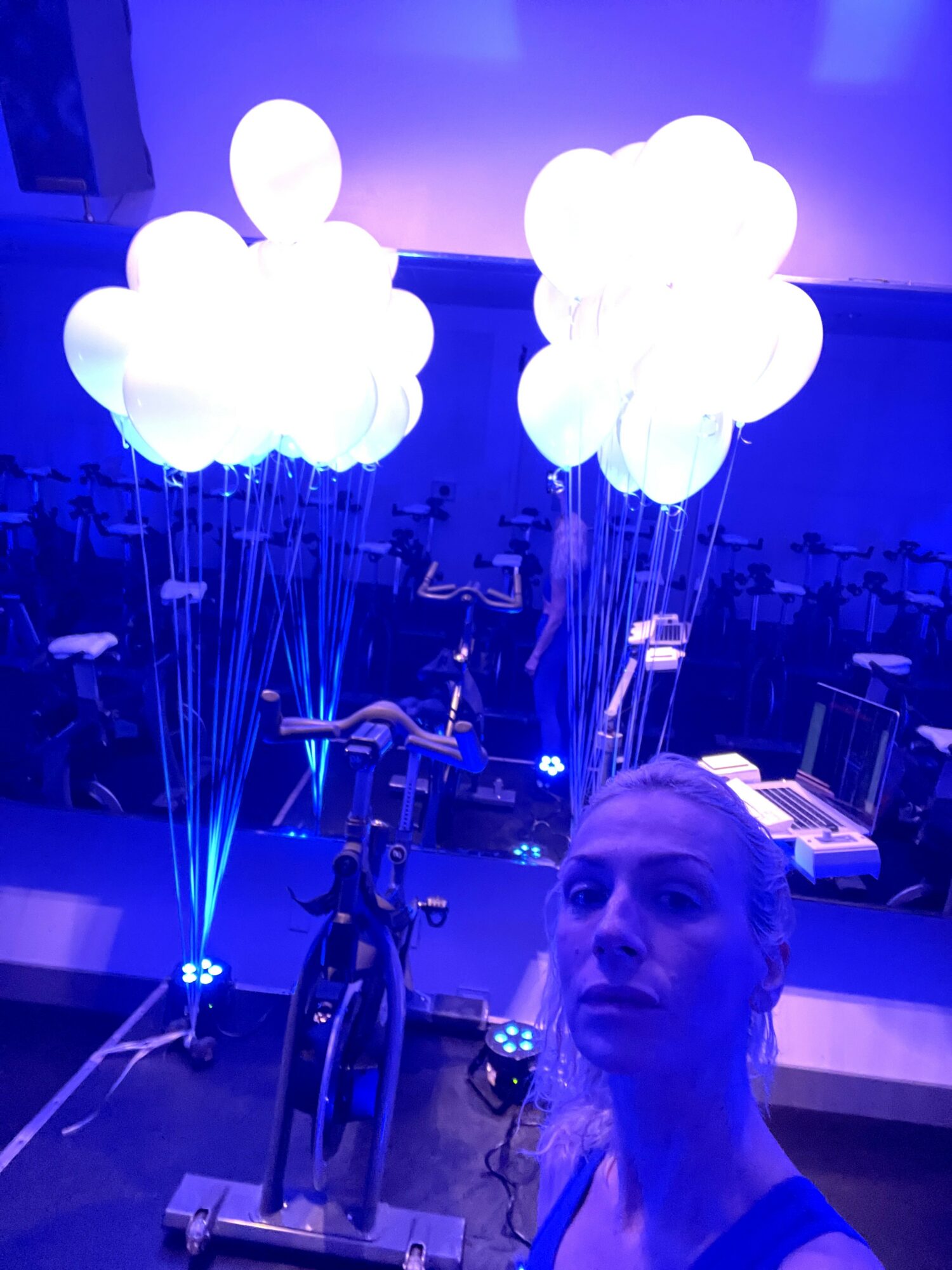 Person taking a selfie in a room with illuminated cloud-shaped decorations and exercise bikes, blue lighting, and tables in the background.